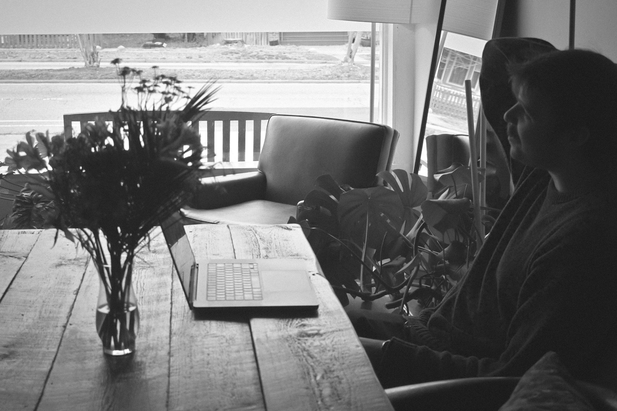 Inside a room, a woman wearing glasses is sitting at a table working on a laptop computer. There are plants on the table, and large windows behind her reveal a sidewalk and street outside.