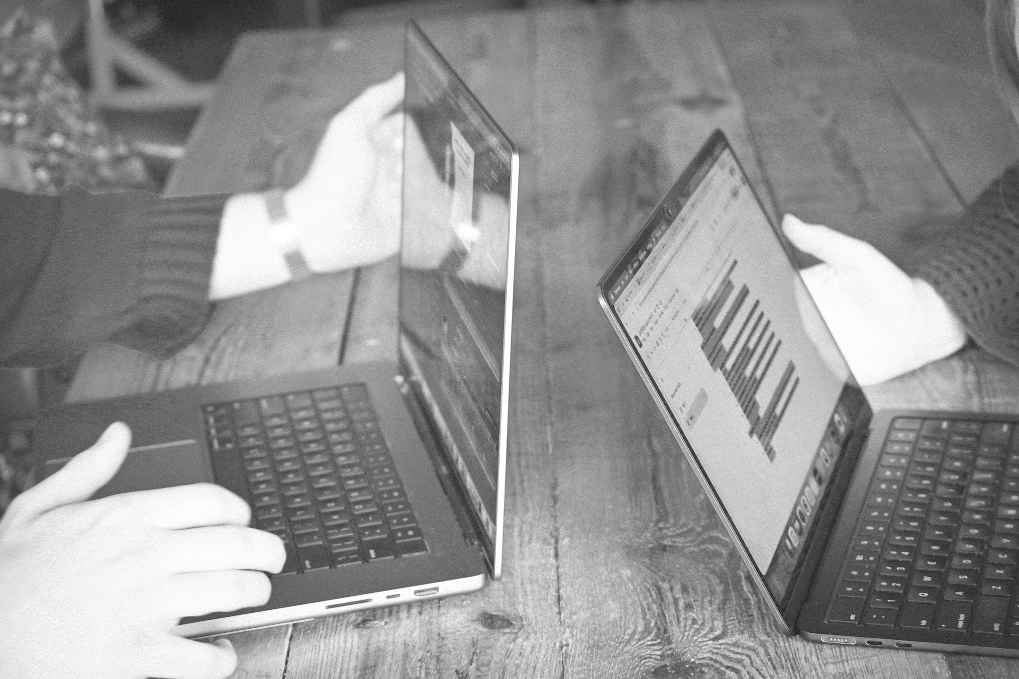 Two people are using laptops on a table, with a mirror reflecting their screens and hands as they type.