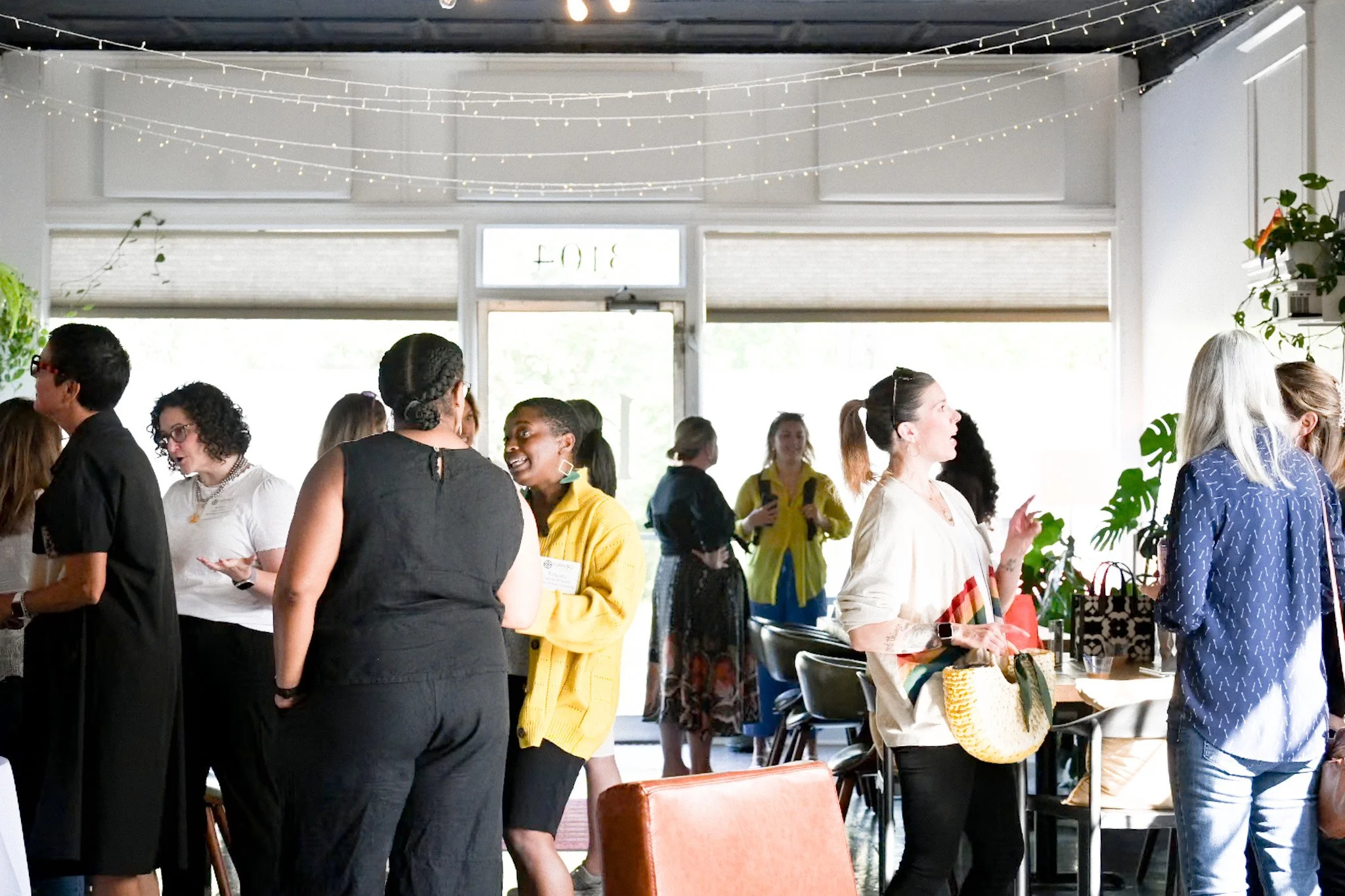 A group of diverse women at a social event or conference, engaging in conversations in a well-lit indoor space decorated with string lights and plants.