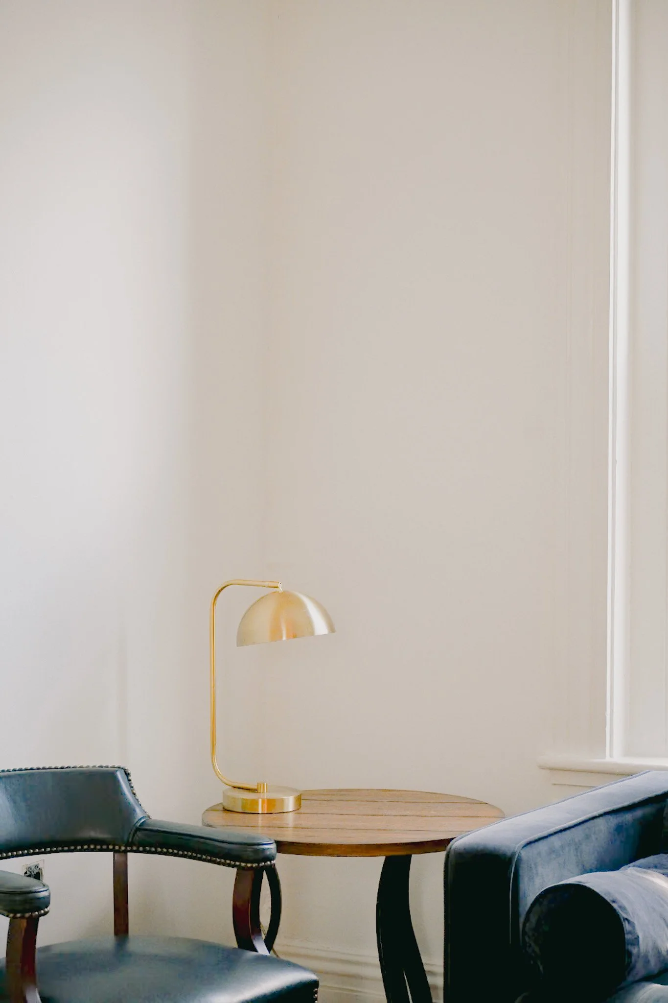 A living room corner featuring a round wooden table, black and gray armchairs, a gold arc floor lamp, and a plain off-white wall.
