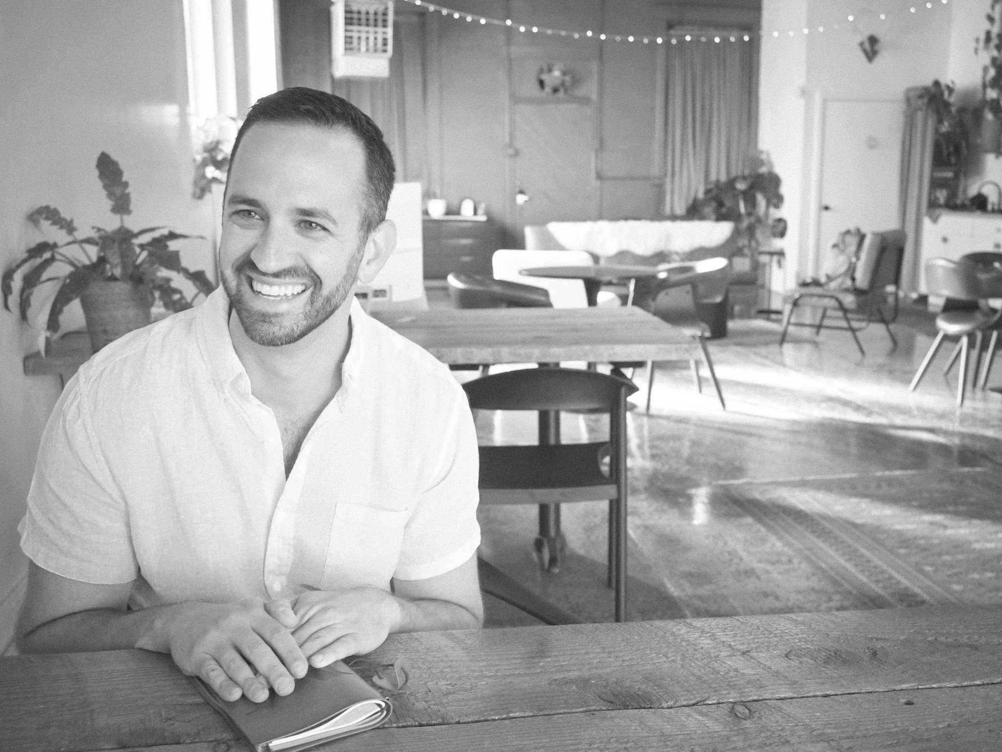 A smiling man with short dark hair and a beard, wearing a light-colored shirt, is sitting at a wooden table in a well-lit room. The room has a casual, cozy atmosphere with bluffs and string lights hanging from the ceiling, and various furniture and decor in the background.