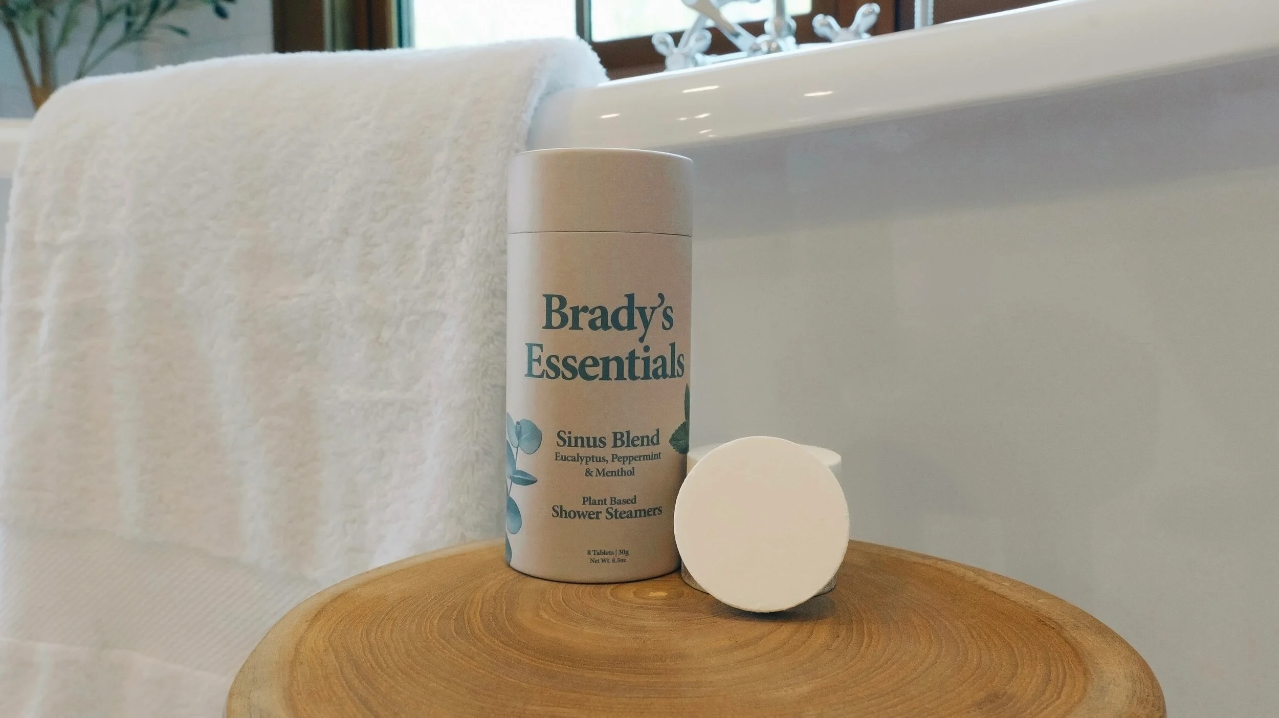 A bottle of Brady's Essentials Sinus Blend plant-based shower steamers with eucalyptus, peppermint, and menthol on a round wooden surface next to a white towel and an egg-shaped soap in a bathroom setting.