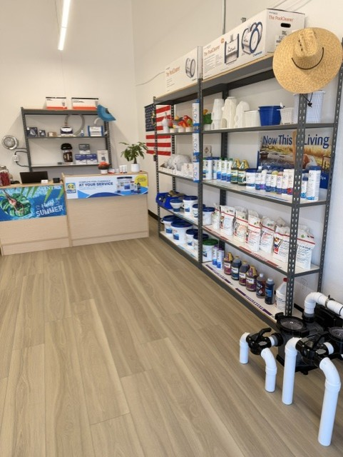 Interior of a retail store with shelves stocked with pool chemicals and equipment, a clipboard on a counter, and a straw hat hanging on the wall.