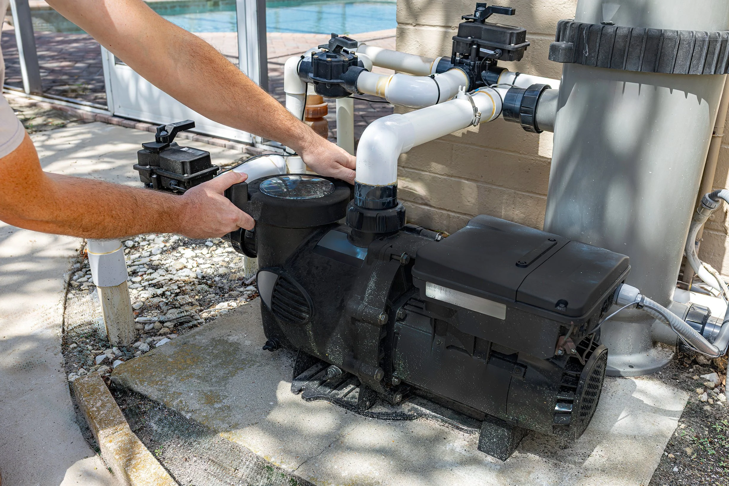 A person working on a pool pump system outdoors, with white PVC pipes and black equipment, next to a swimming pool and brick patio.