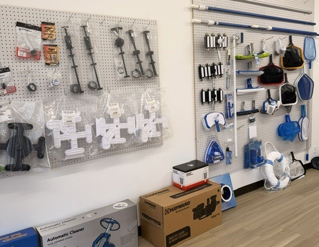 Display of vacuum cleaner parts and accessories on pegboards in a store or workshop, including hoses, brush attachments, and filters, with boxed vacuum cleaners on the floor.