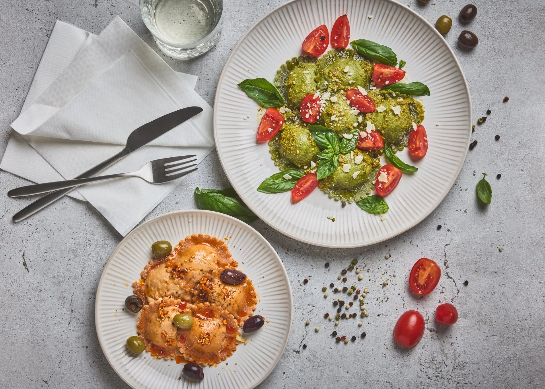 Ein Teller mit gekochten Nudeln, Kirschtomaten, Basilikumblättern und Parmesan, daneben ein Glas Wasser und eine Serviette mit Besteck.