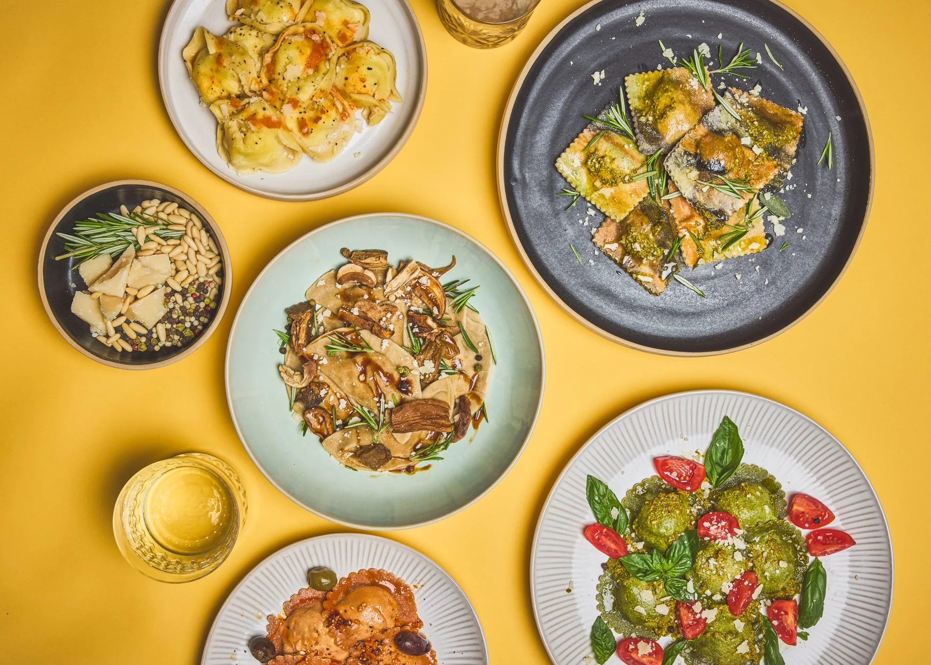 Bunte italienische Gerichte auf mehreren Tellern, darunter Pasta, Salat, Pilz-Pasta und Aufschnitt, alles auf einem gelben Tisch.