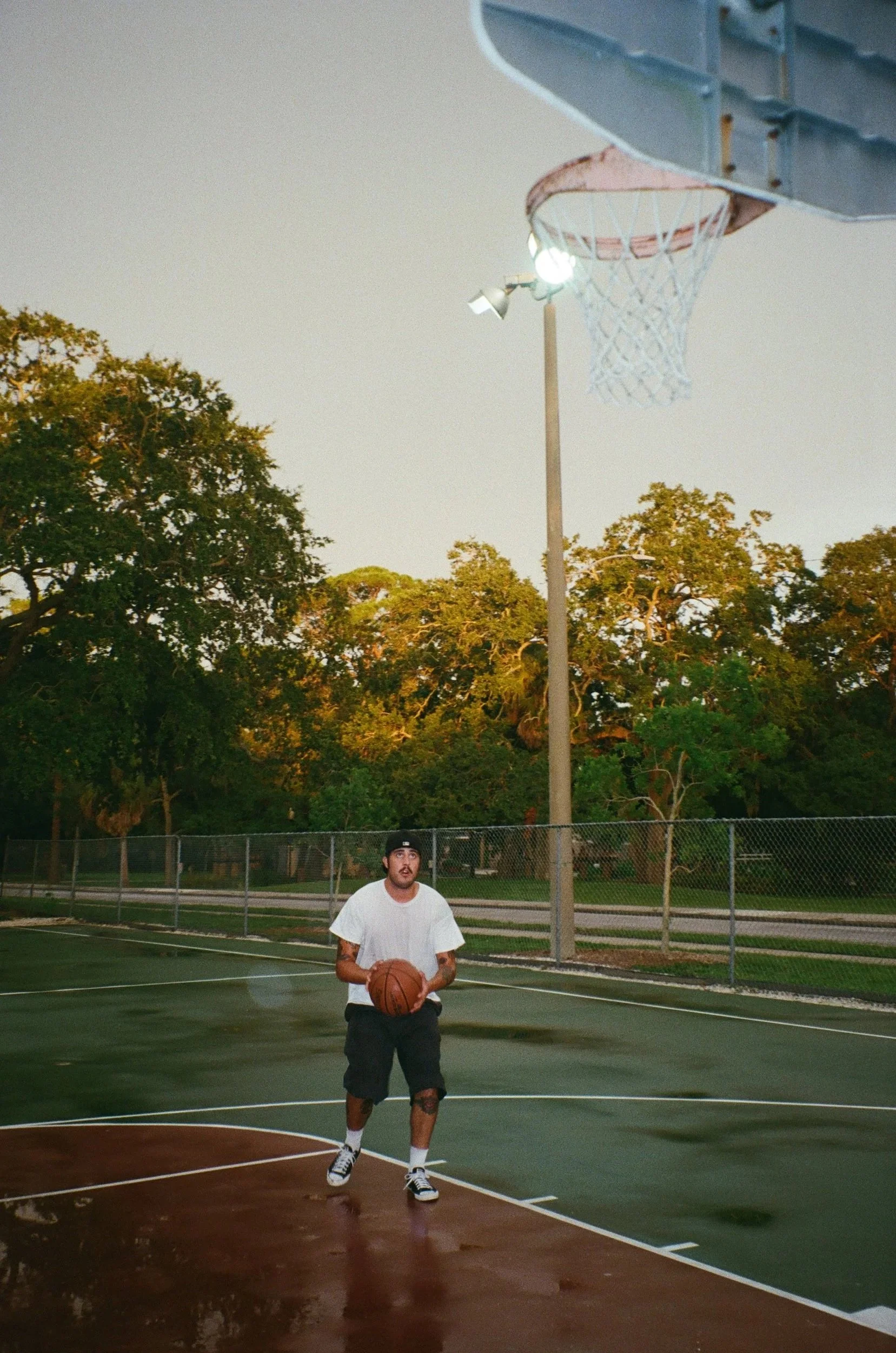 A man with a beard, tattoos, and short hair, wearing a white t-shirt and black shorts, holding a basketball on an outdoor basketball court during sunset, with trees and a chain-link fence in the background.