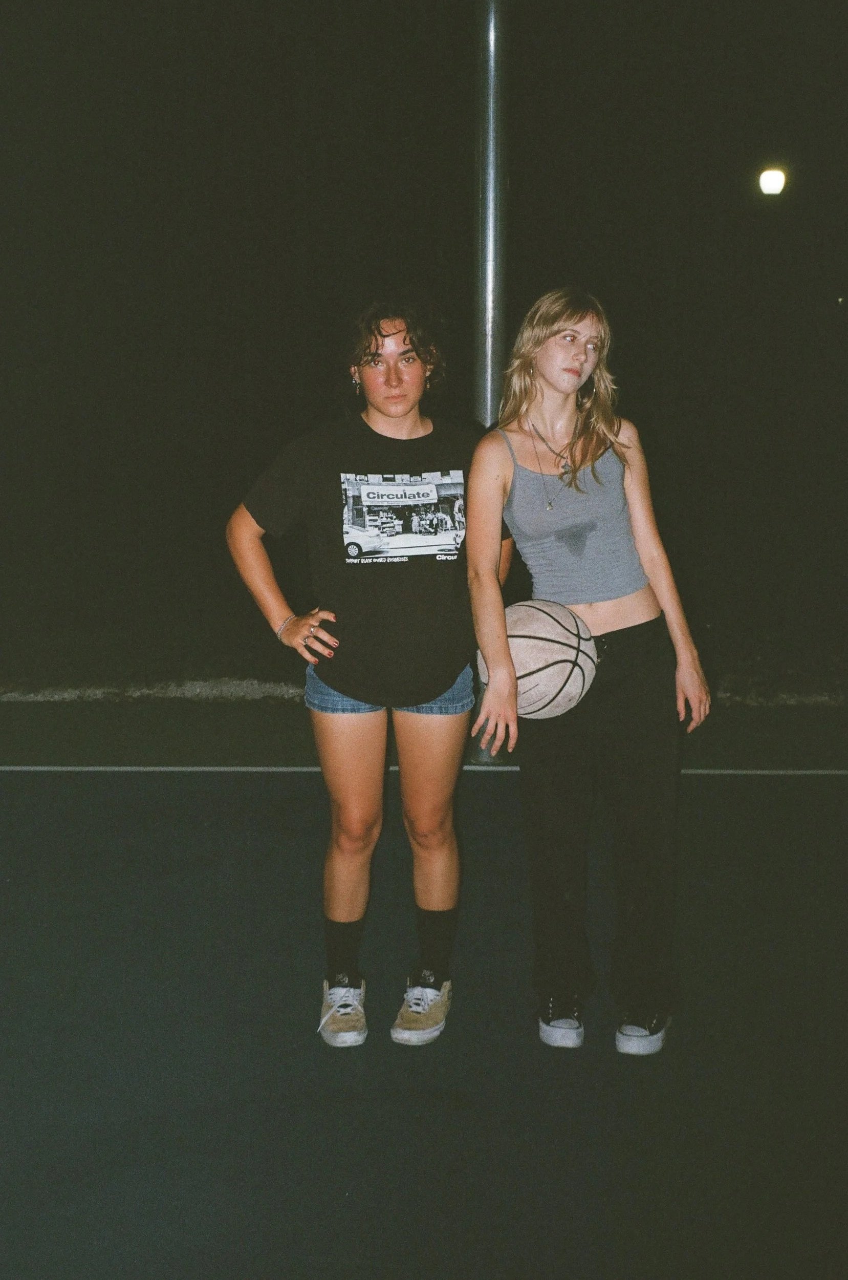 Two young women standing on a basketball court at night, with one holding a basketball, both dressed casually in tank tops, shorts, and sneakers.