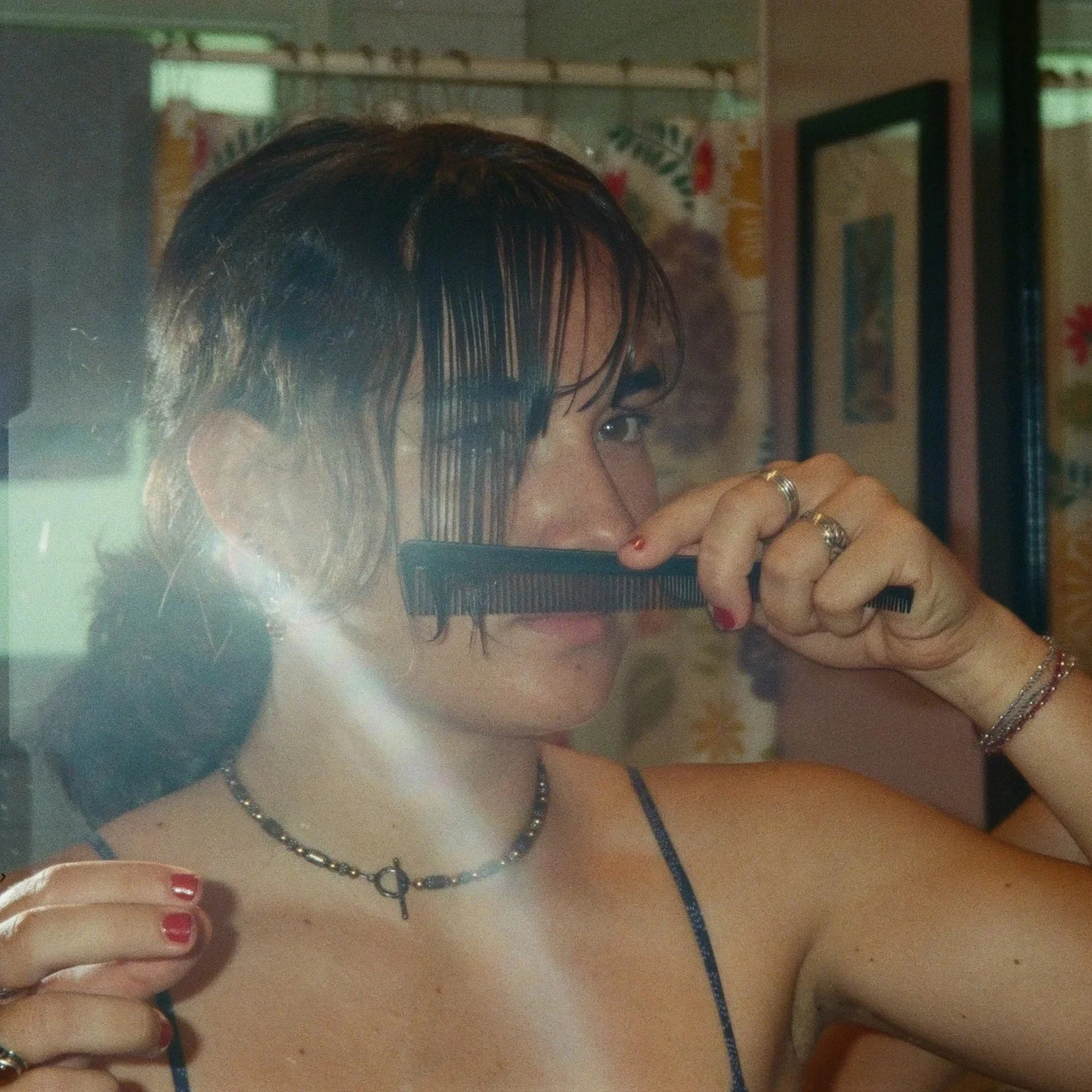 A woman with wet hair, wearing rings, a necklace, and red nail polish, holds a comb near her face and takes a mirror selfie in a room with framed pictures and patterned wallpaper in the background.