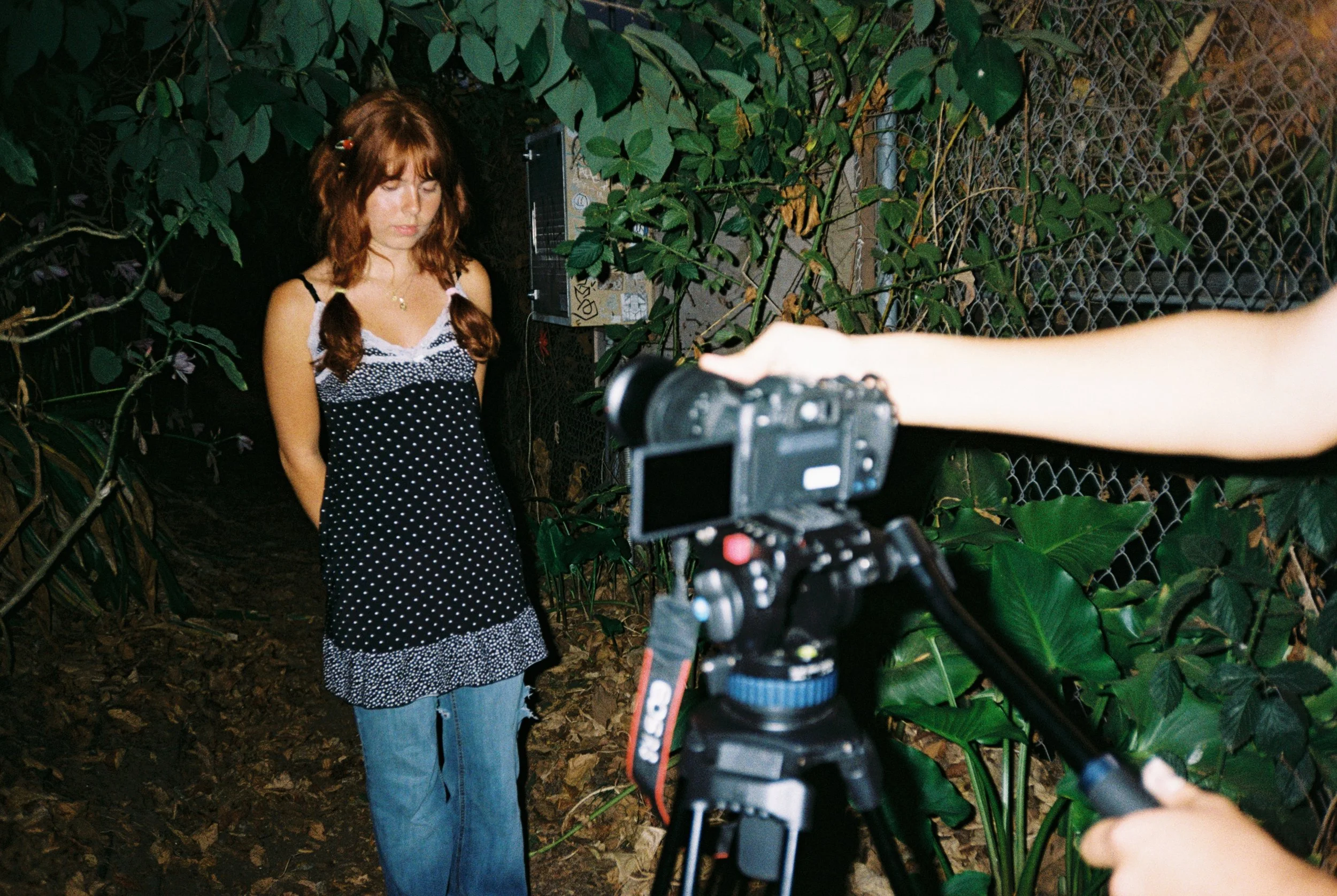A woman with red hair styled in pigtails, wearing a black and white polka dot dress and jeans, stands outdoors near a chain-link fence and green foliage. A person holding a professional camera is taking her photograph at night.
