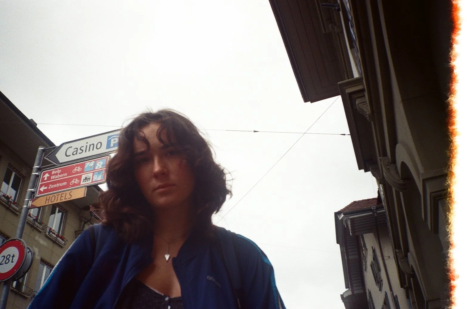 A young woman with dark, curly hair standing outdoors, with signs indicating directions to a casino, hotels, and various city areas in the background.