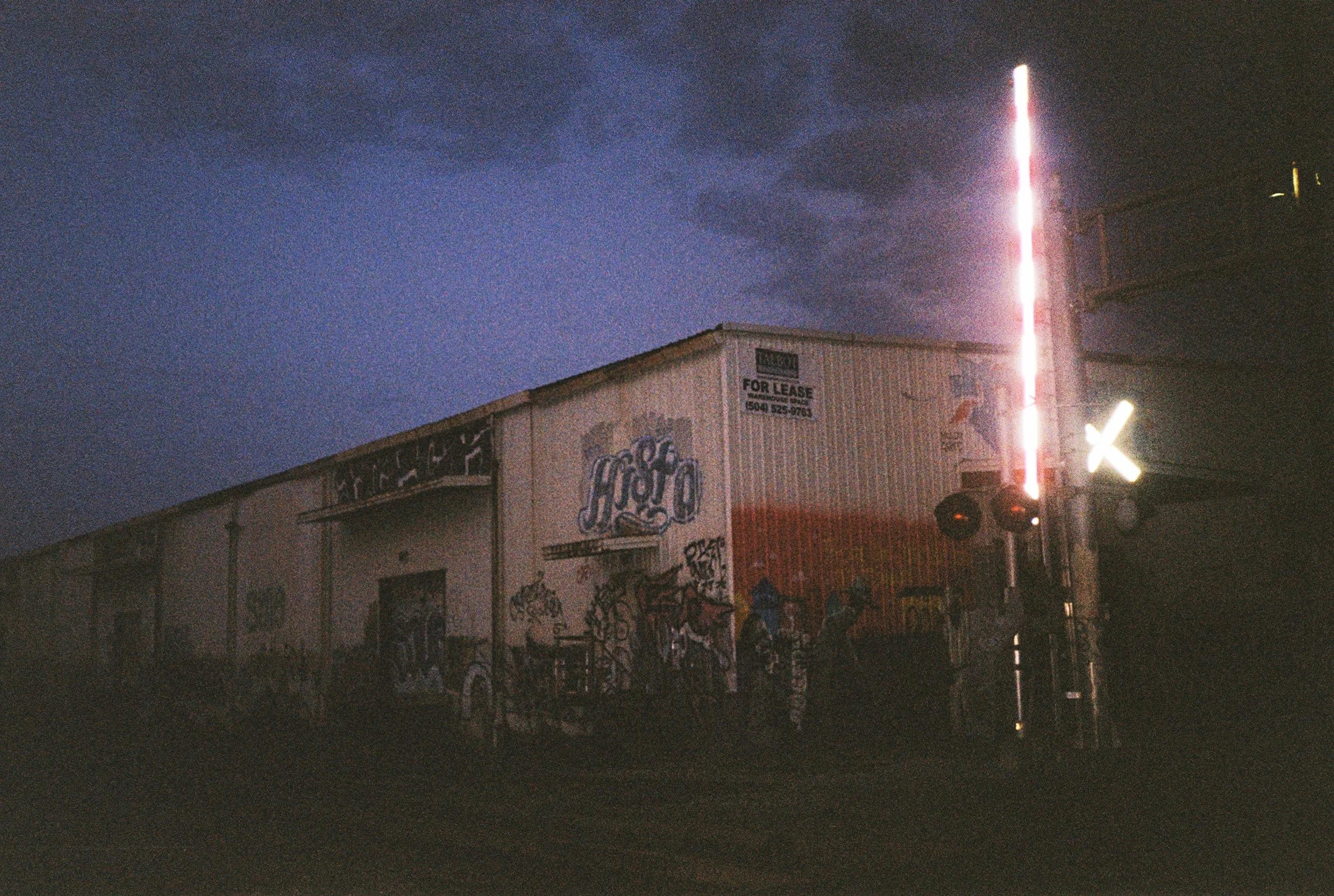 Street scene at night with a building covered in graffiti, a 'For Lease' sign, and bright lights, with dark clouds in the sky above.