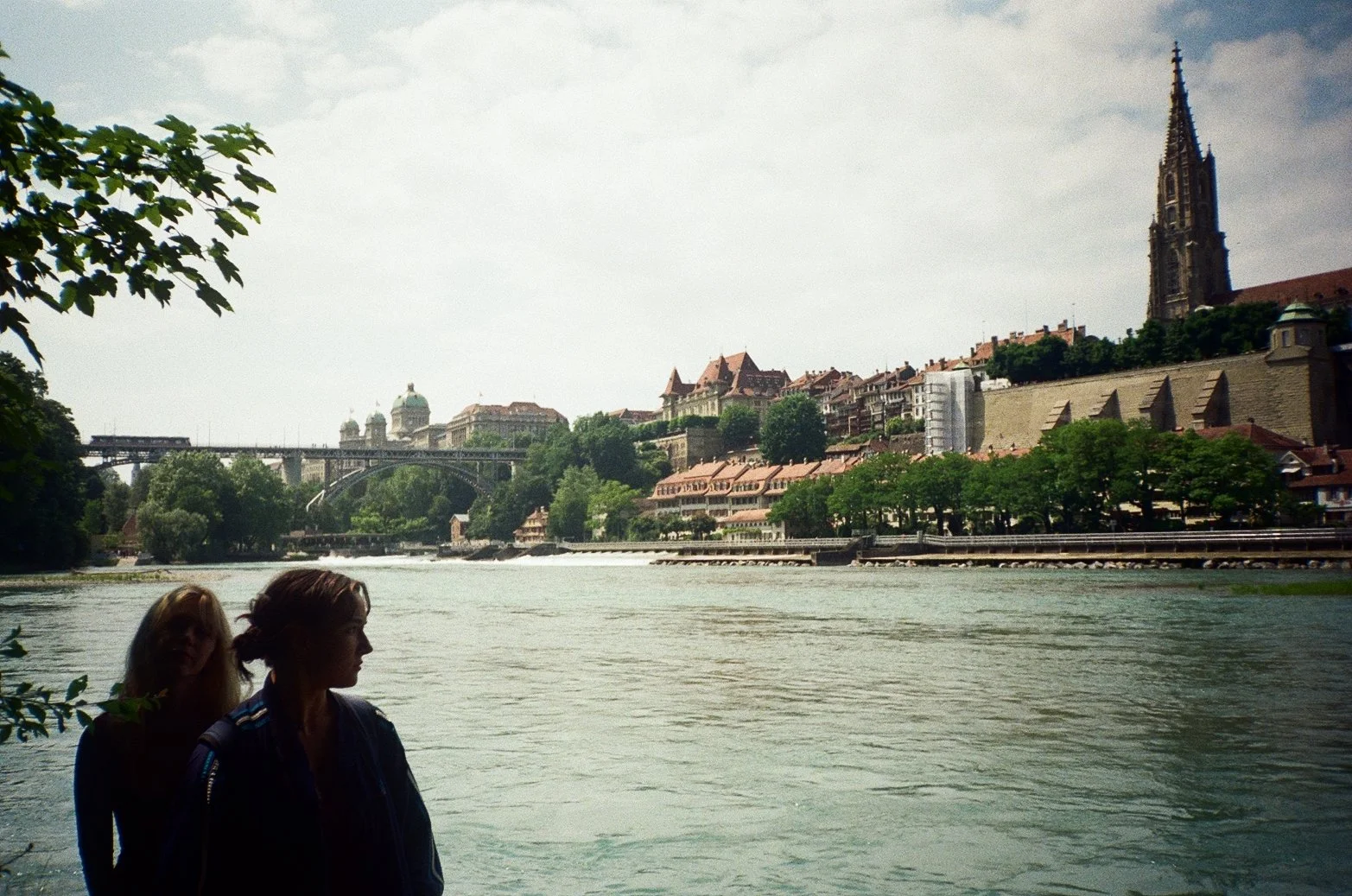 People standing by a river with historic buildings, a bridge, and a church with a tall steeple in the background.