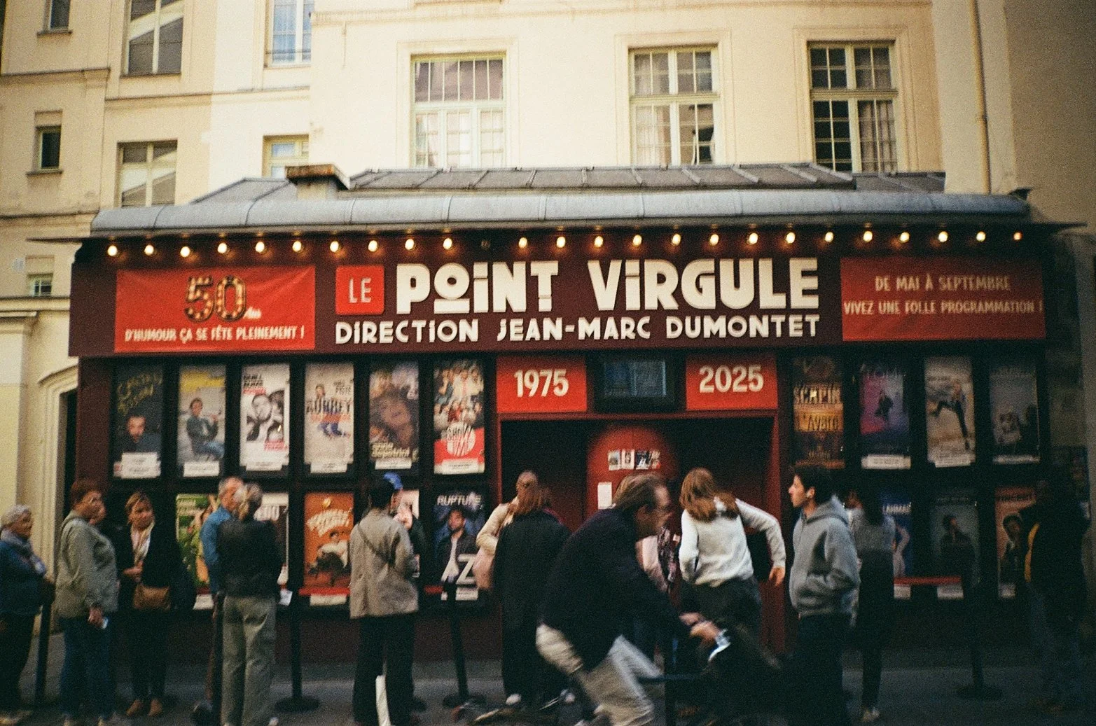 A theater marquee celebrating 50 years of Le Point Virgule, a comedy venue, with posters of performances and patrons waiting outside.