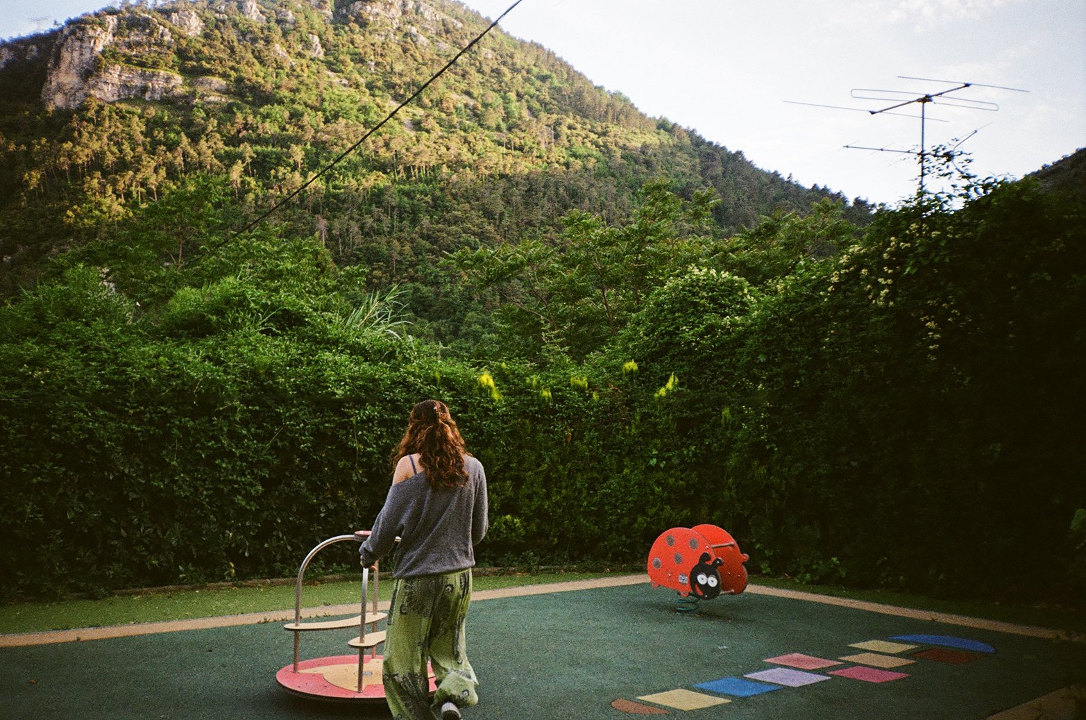 A person with long curly hair, wearing a gray sweater and patterned pants, standing on a children's playground with a ladybug-shaped spring rocker and colorful hopscotch markings, surrounded by green bushes and trees with a mountain in the background