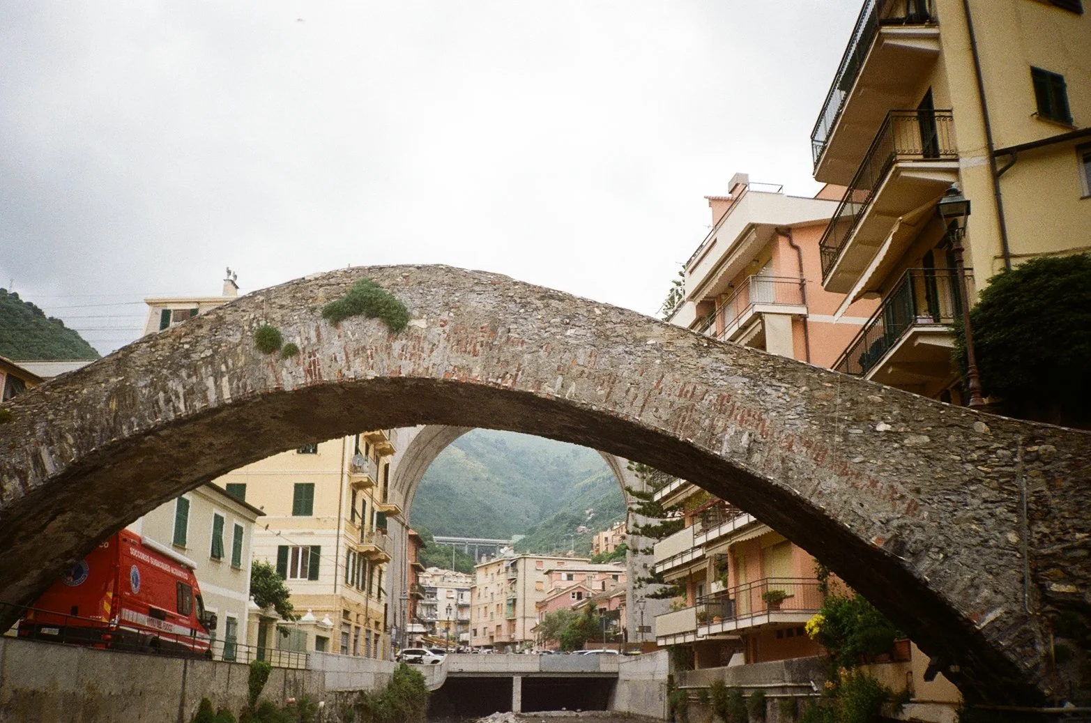 A stone arch bridge over a small urban canal with buildings on both sides, set against a mountainous background and cloudy sky.