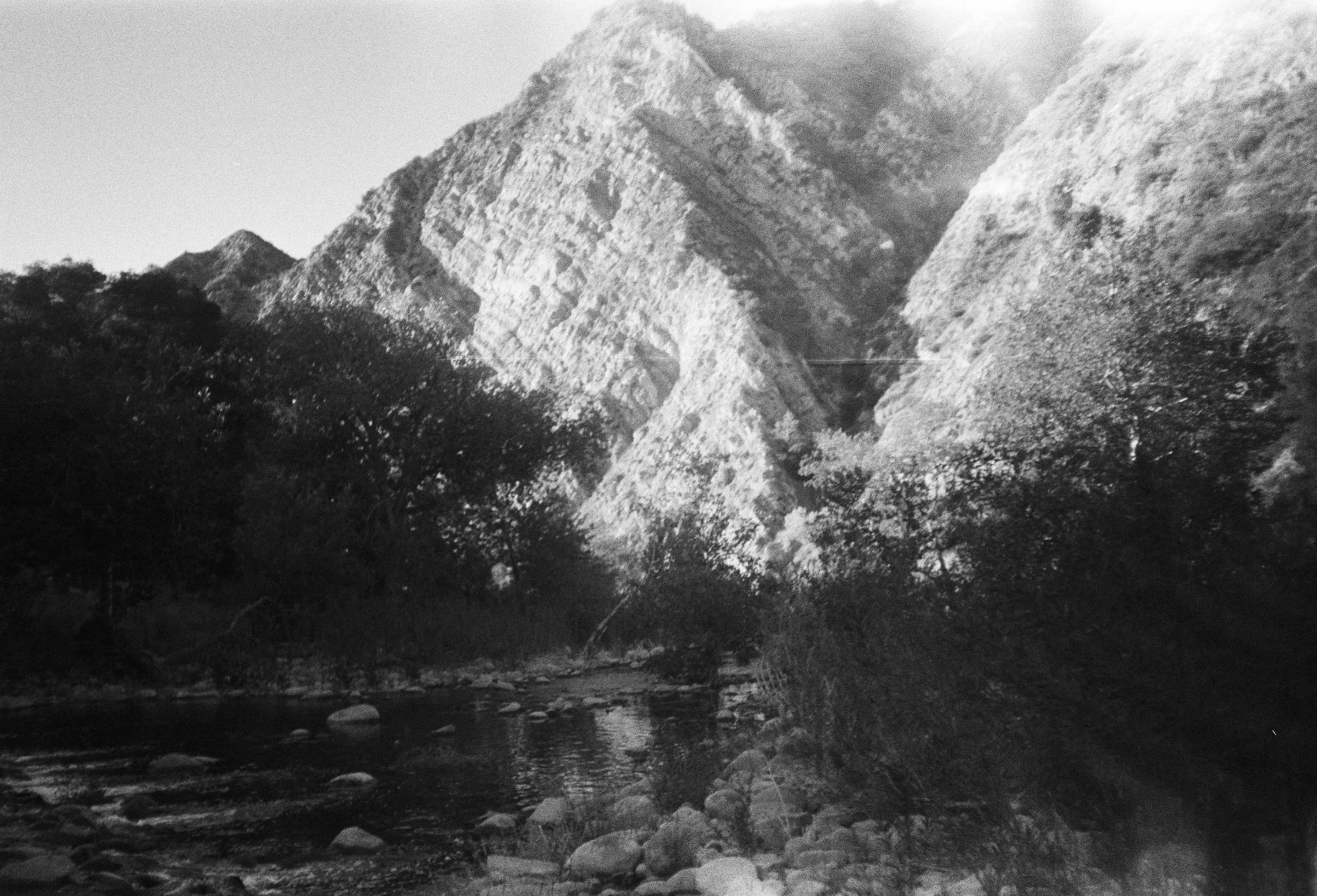 Black and white photo of rugged mountains with layered rock formations, a treeline at the base, and a rocky river or stream in the foreground.