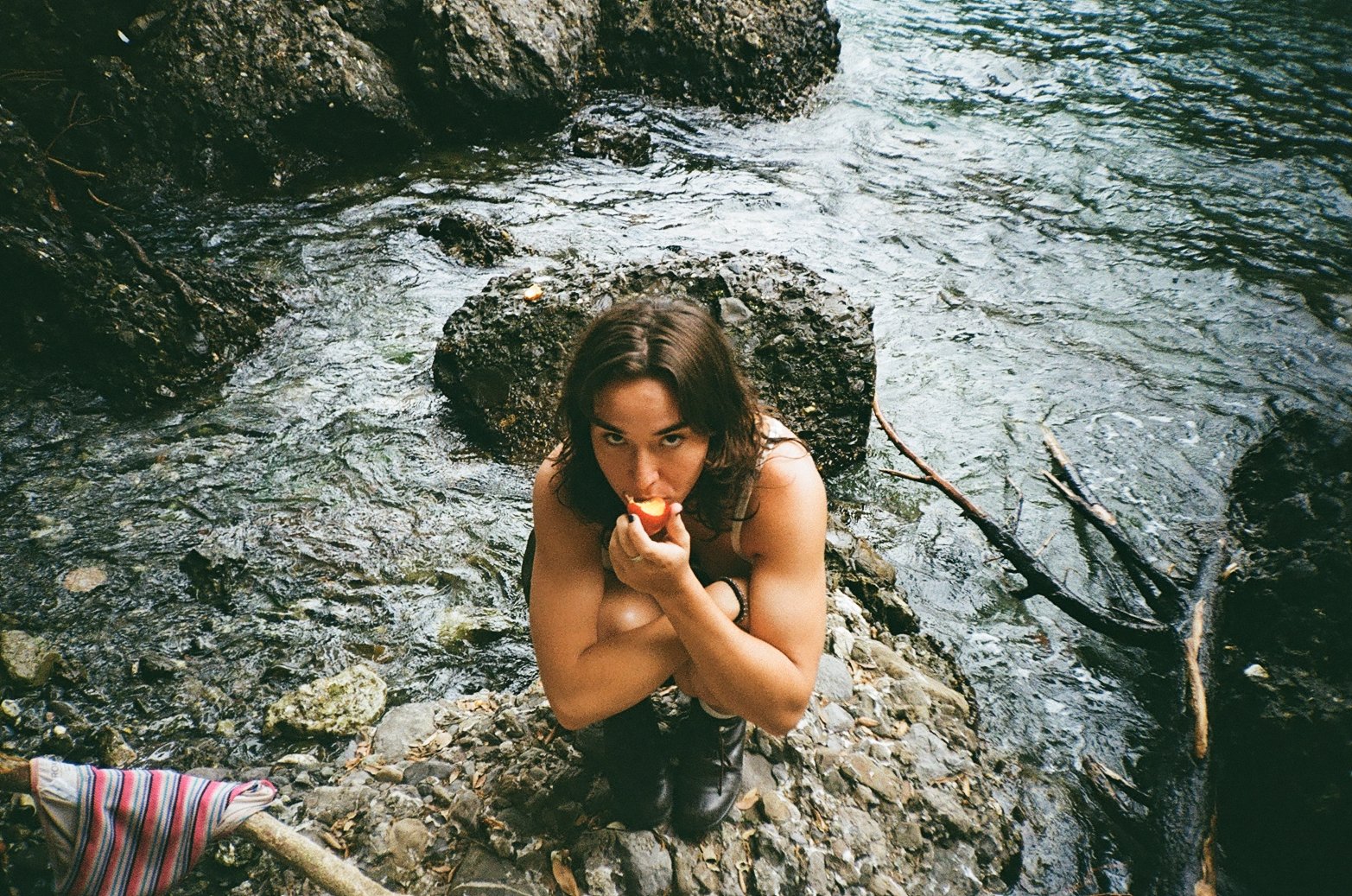 Woman with dark hair and bands on her arm, crouching on rocky terrain near a stream, eating an apple.