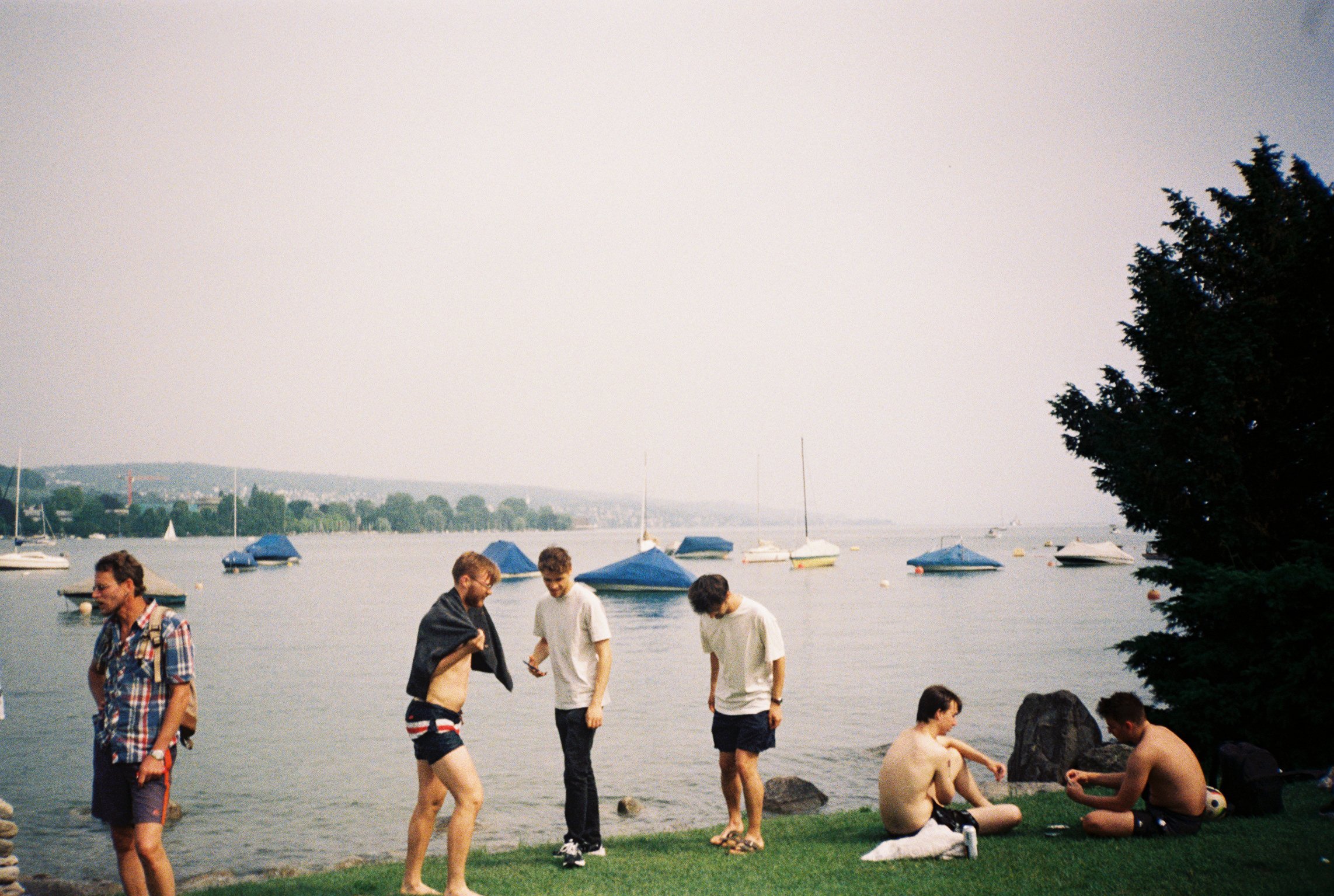 People gather near a lakeside on a cloudy day, with boats anchored on the water and a tree to the right, and a distant shoreline with houses and trees.