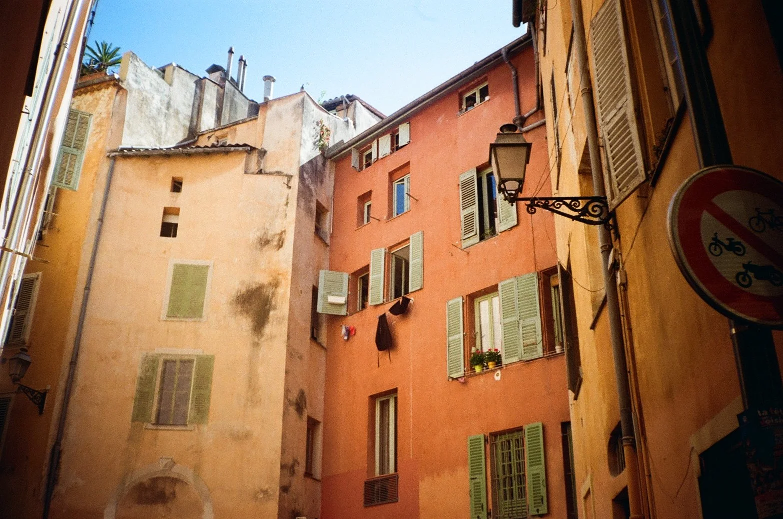 Narrow European street with tall, colorful buildings featuring shuttered windows and floral window boxes. A street lamp and a street sign are visible.