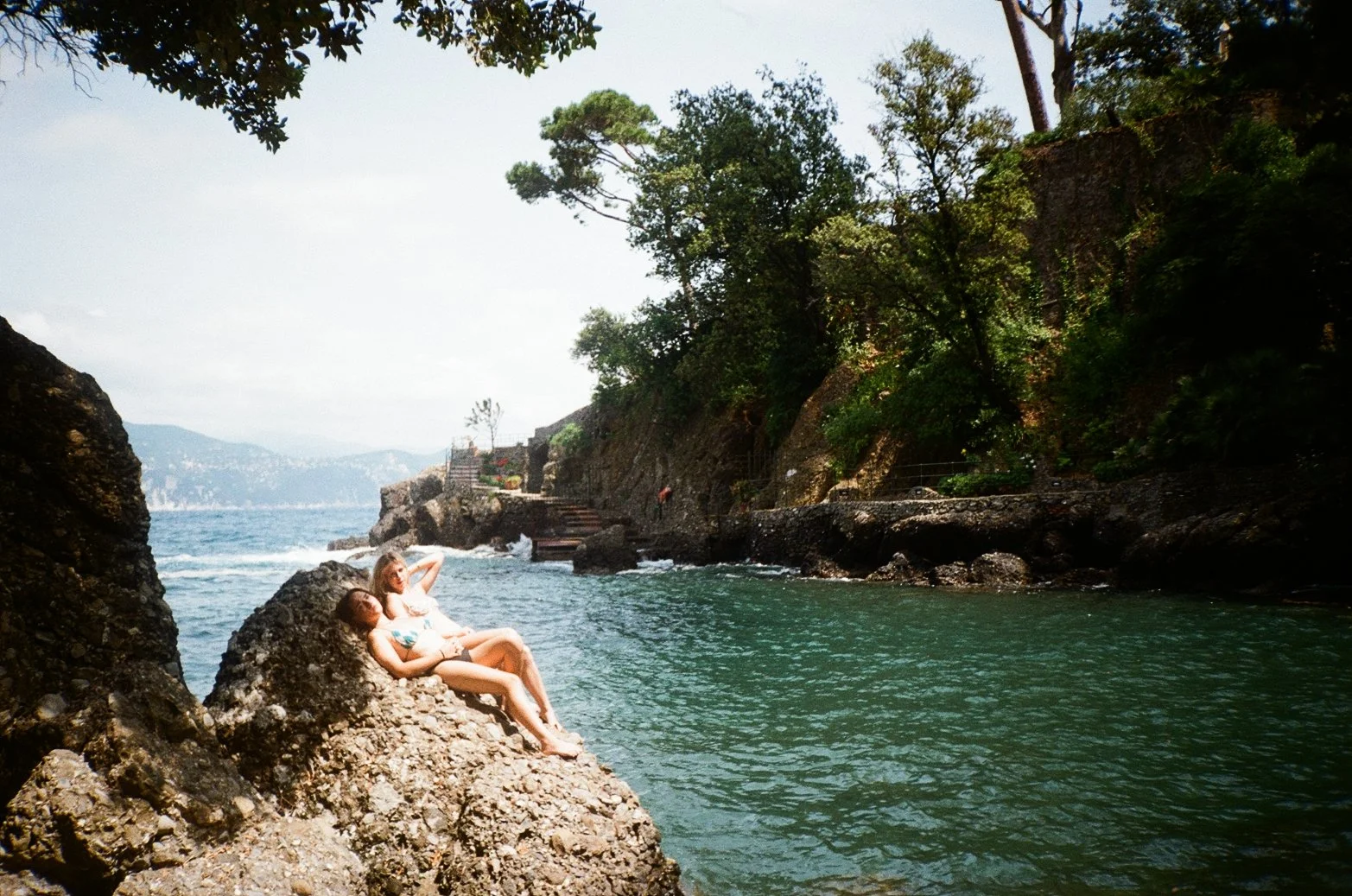 Two women relaxing on rocks by the sea with greenery and cliffs in the background on a sunny day.