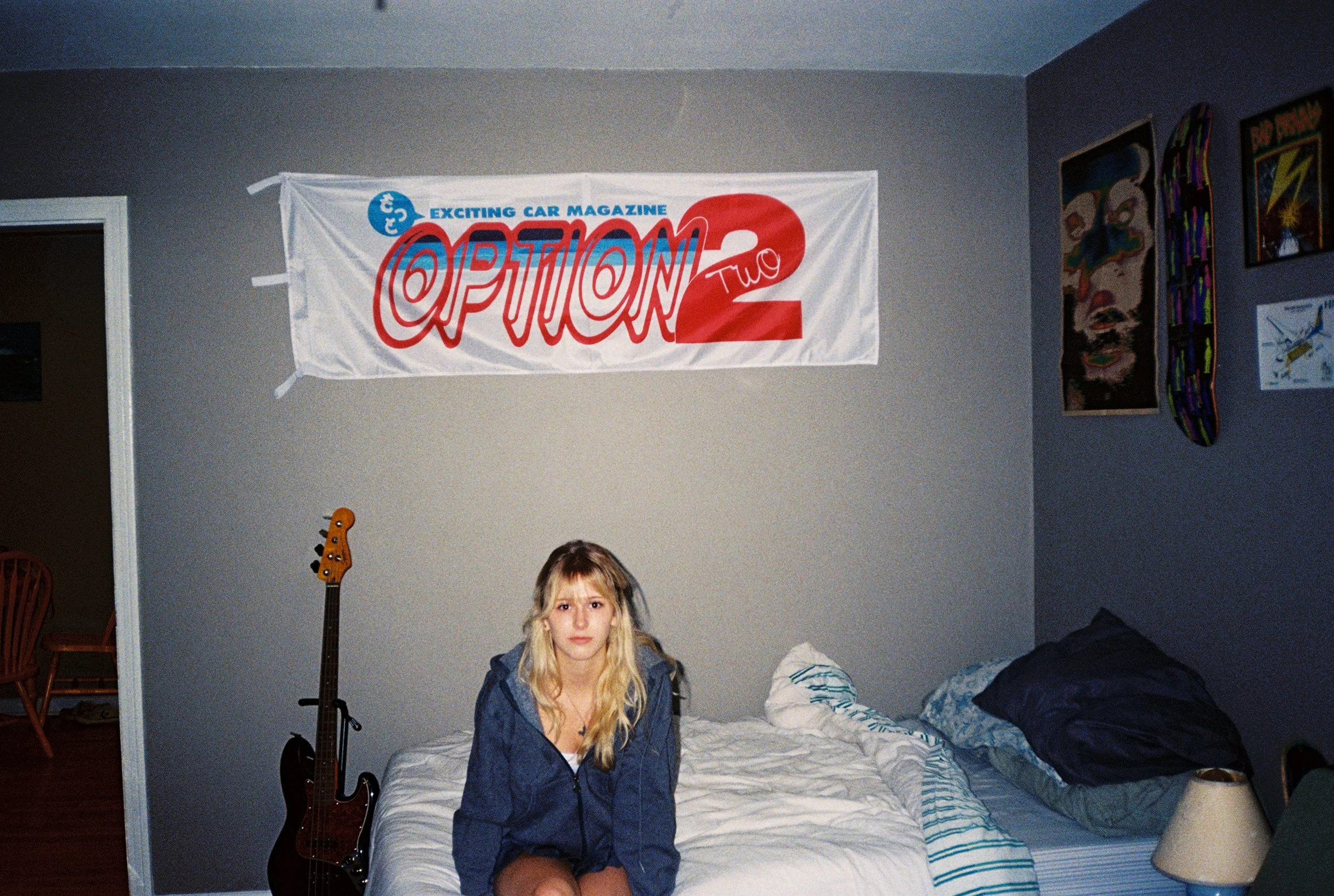 A young woman with blonde hair sitting on a bed with white sheets and striped pillowcases, wearing a dark blue jacket. Behind her, a wall with artwork and a banner that reads 'Exciting Car Magazine Option 2'. A guitar stands beside her, and a person 