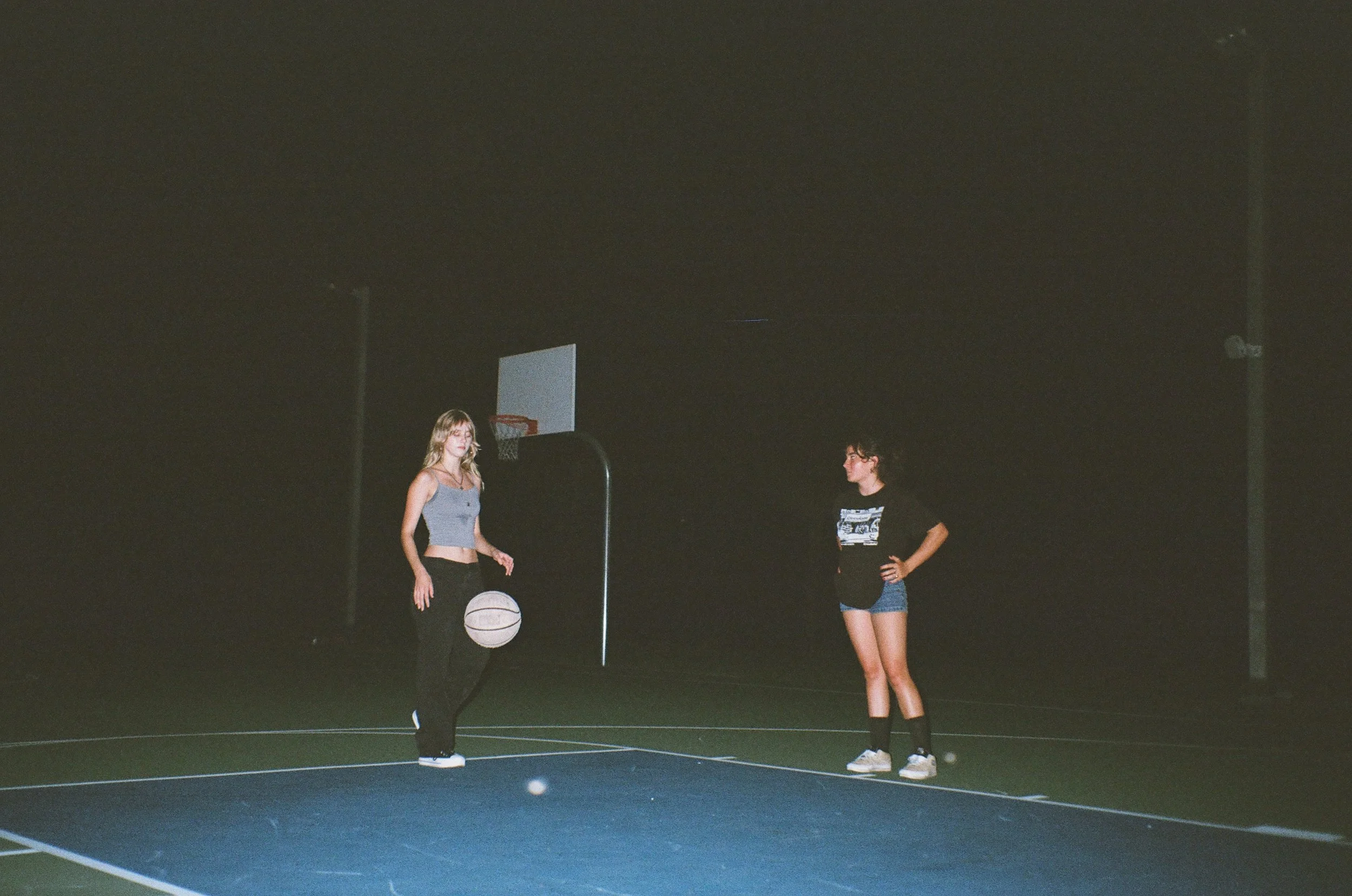 Two young women standing on a dark outdoor basketball court at night, with one woman preparing to shoot a basketball towards the hoop.