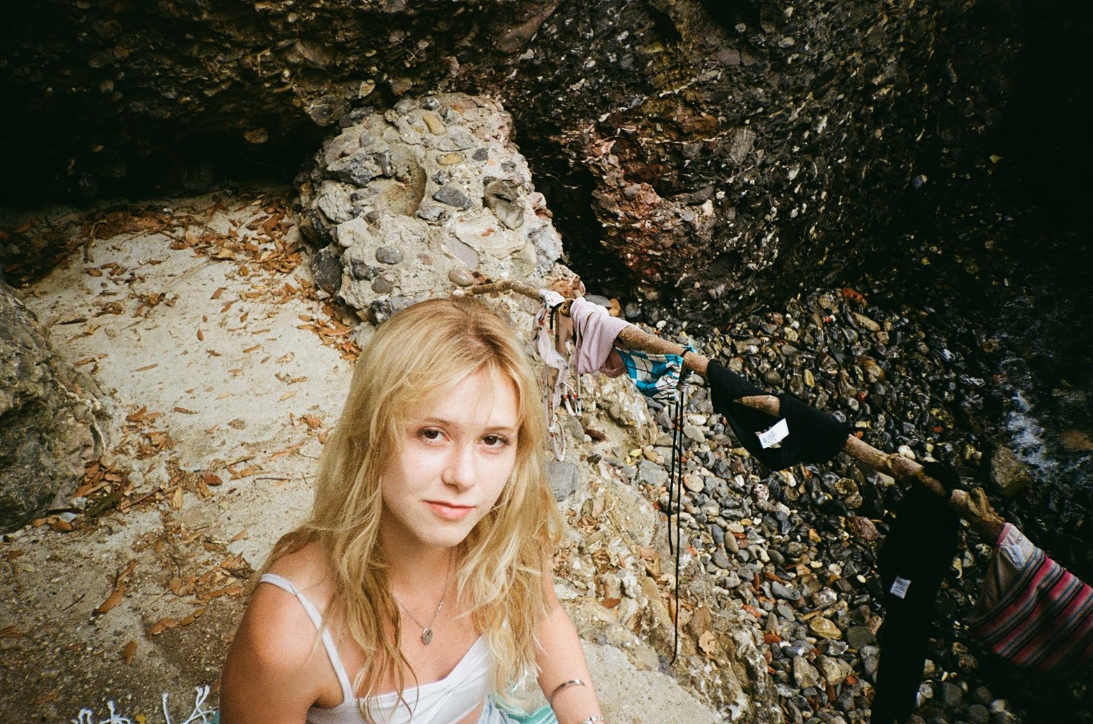 A young woman with blonde hair and a white tank top sitting on a sandy and rocky beach, with a rocky cliff behind her and a rope tied to a stick next to her, extending into the water.