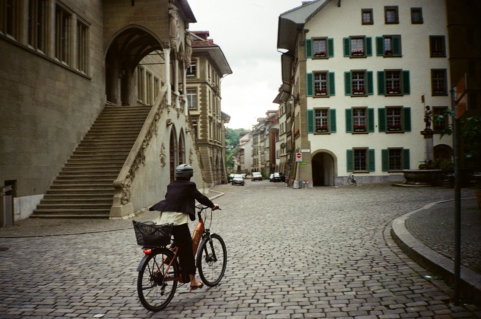 A woman riding a bicycle on a cobblestone street in a European city with historic buildings and colorful window shutters.