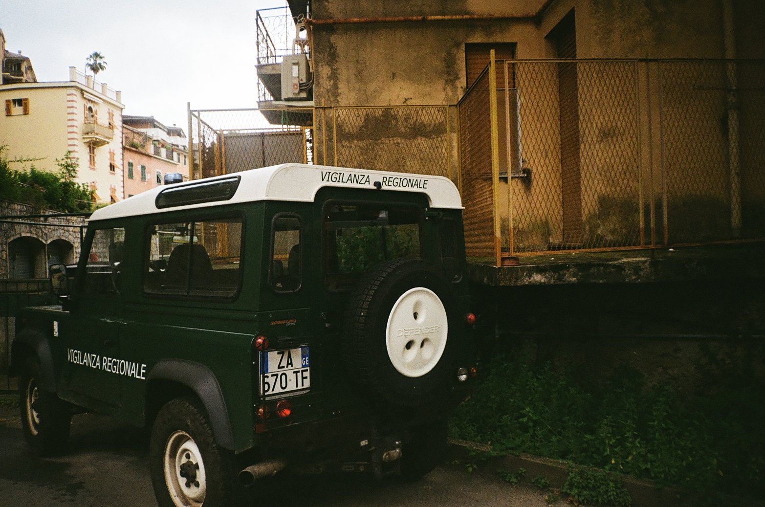 A green police vehicle marked 'Vigilanza Regionale' parked next to a yellow metal fence on a city street, with apartment buildings in the background.