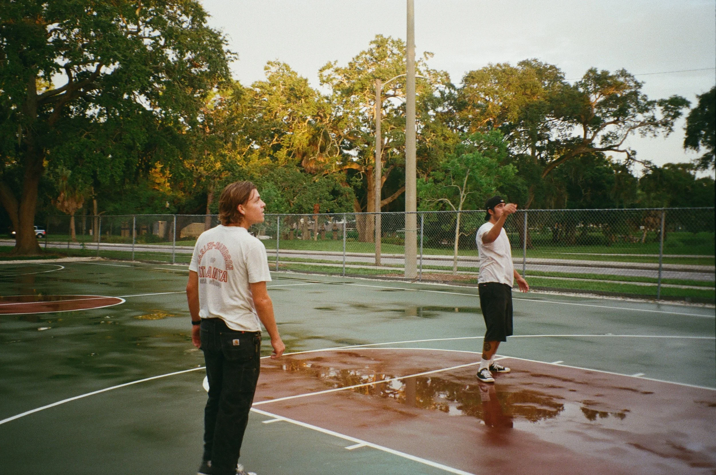Two young men standing on a wet outdoor basketball court with trees in the background. One is wearing a white t-shirt with red lettering and the other has a baseball cap and white t-shirt.