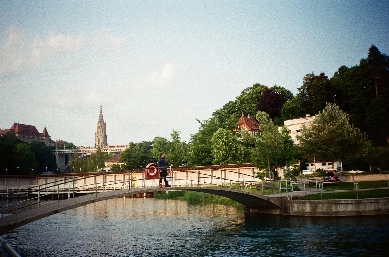A person standing on a small bridge over a river, with lush green trees and buildings in the background, including a tall church spire.
