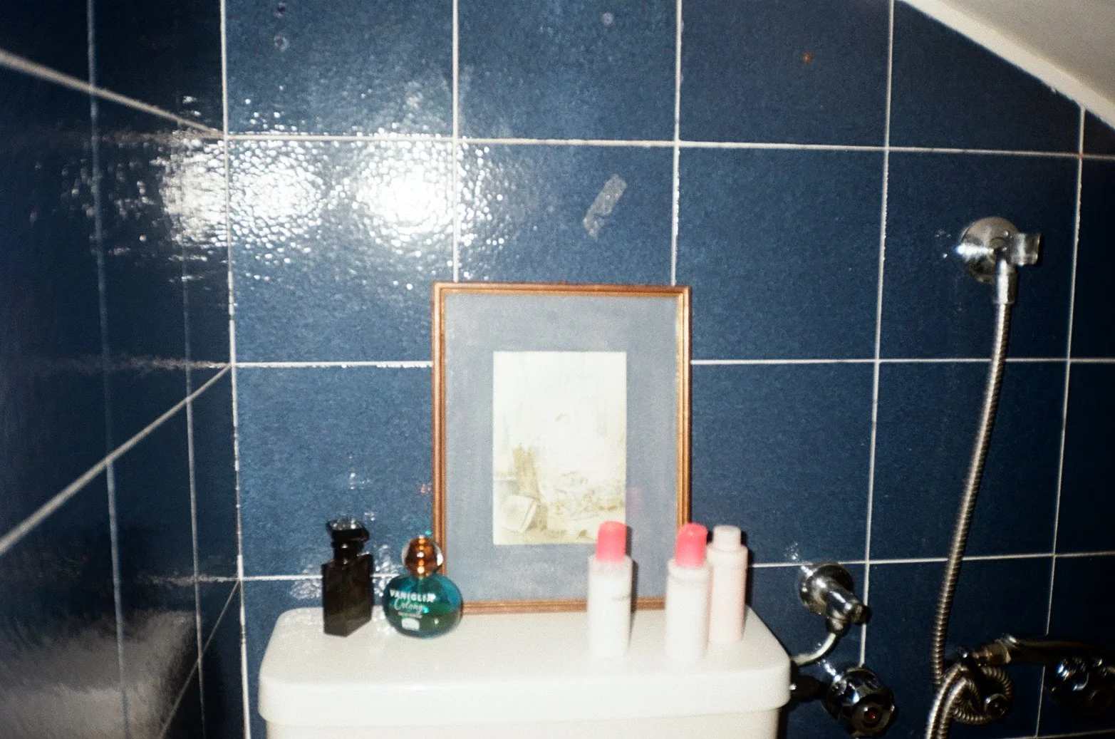 A bathroom with blue tiled walls, a mirror, and various toiletries on top of a white sink or cabinet. There is a handheld shower attached to the wall with a hose.