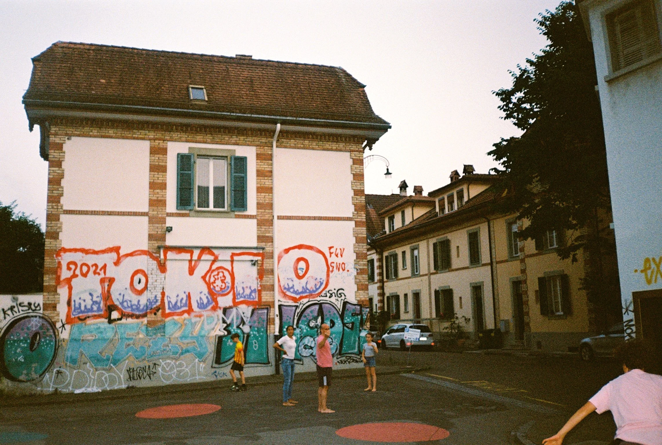 Street scene with graffiti-covered wall, people standing and walking, and residential buildings in the background.