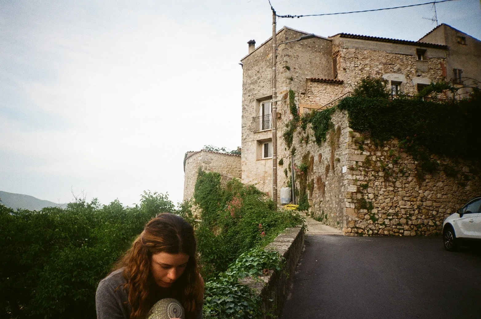 A young woman with long, wavy brown hair looking down, standing in a rural area with an old stone house, greenery, and a car in the background at dusk.