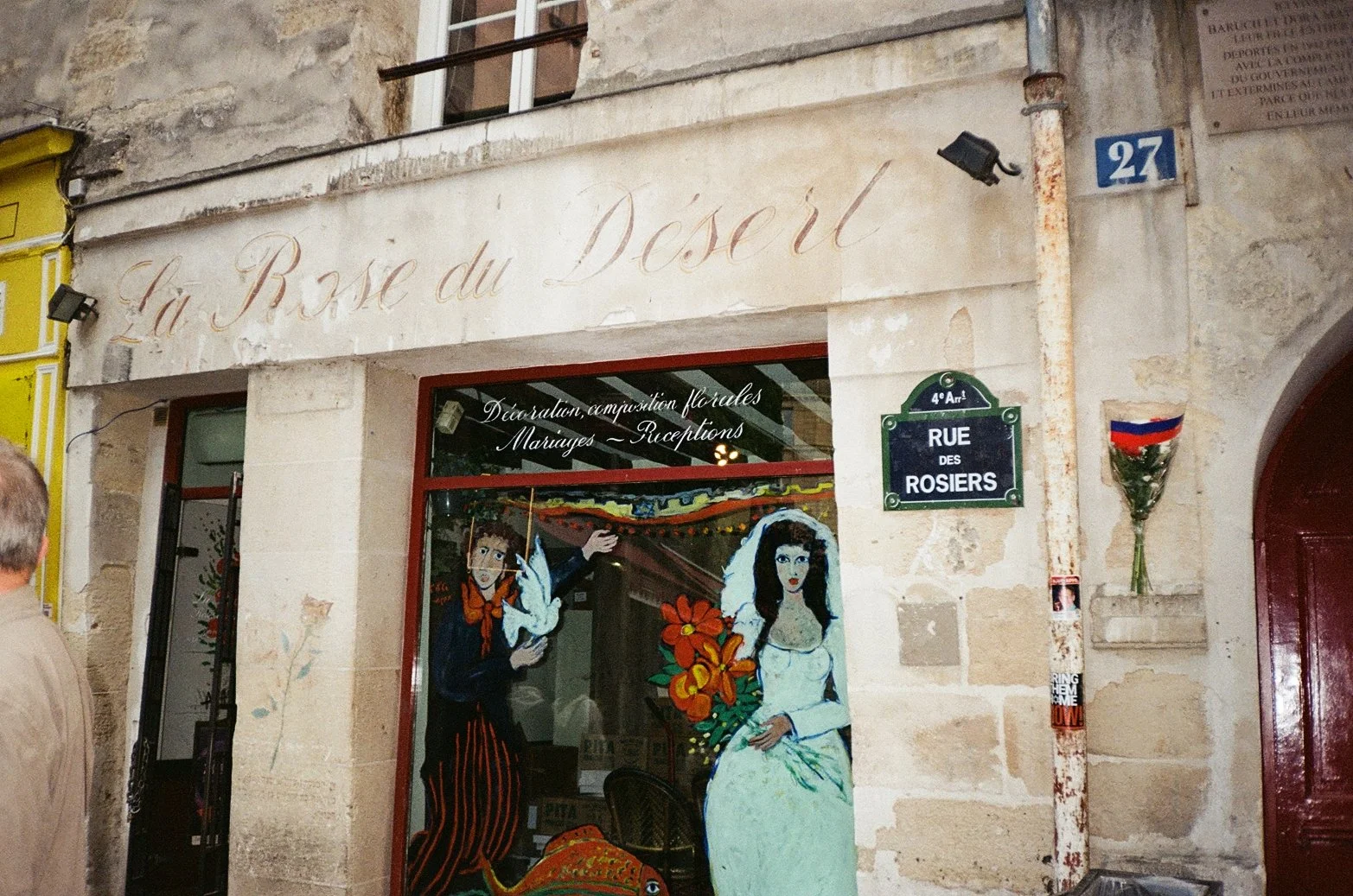 The exterior of a shop on Rue des Rosiers in Paris, with painted illustrations of a woman in a wedding dress and a girl holding a dove in the window. The shop has a sign in French listing services like flower arrangements, weddings, and receptions.