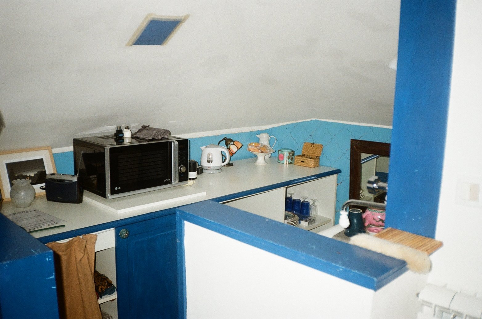 Kitchen countertop with microwave, electric kettle, decorative items, jars, and glassware, with blue and white cabinetry and a wall with a crackled paint pattern.