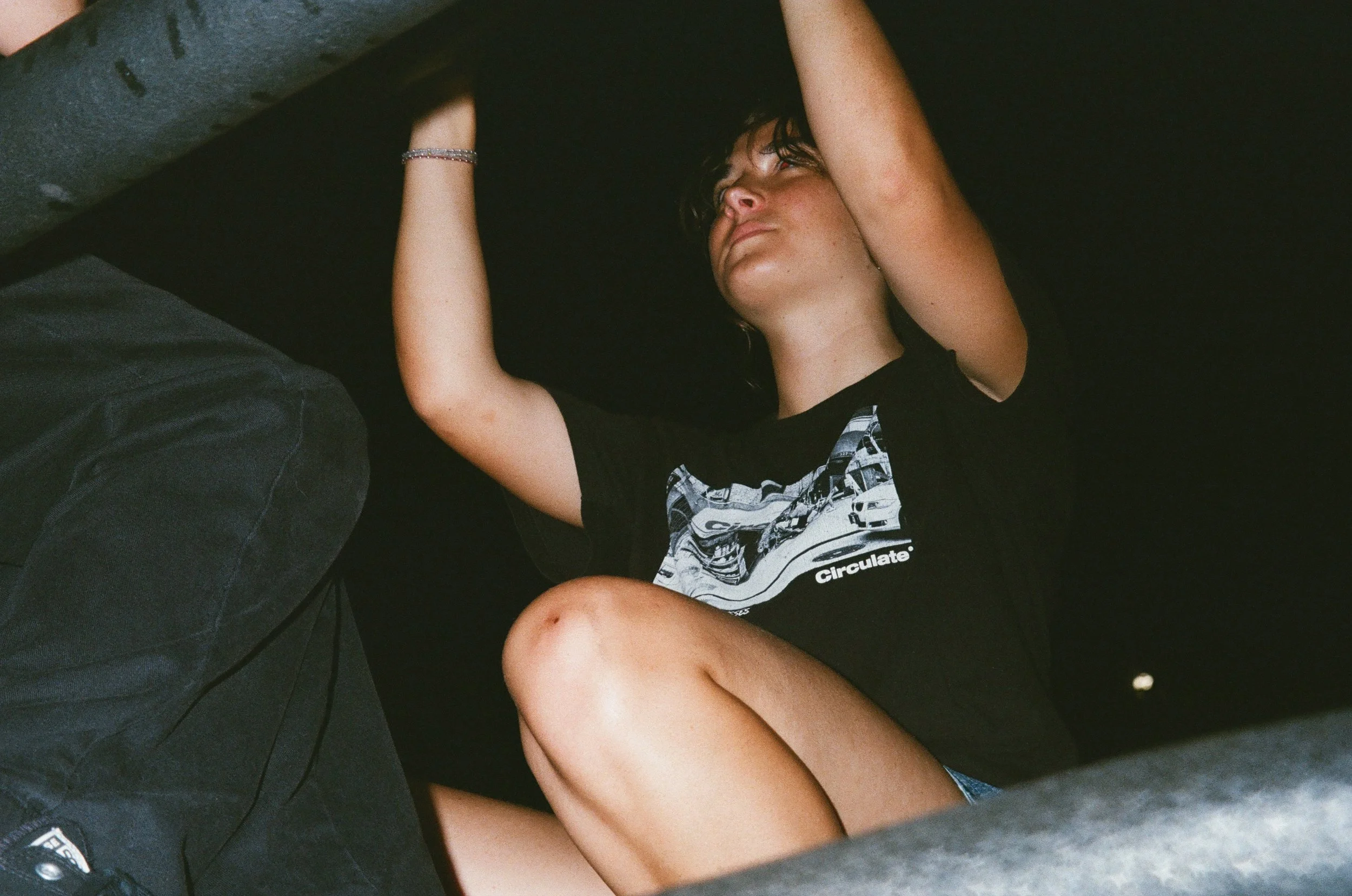 Person lying down with slightly curly dark hair, wearing a black t-shirt with a graphic design, resting on a black surface, looking upward with arms extended above head.