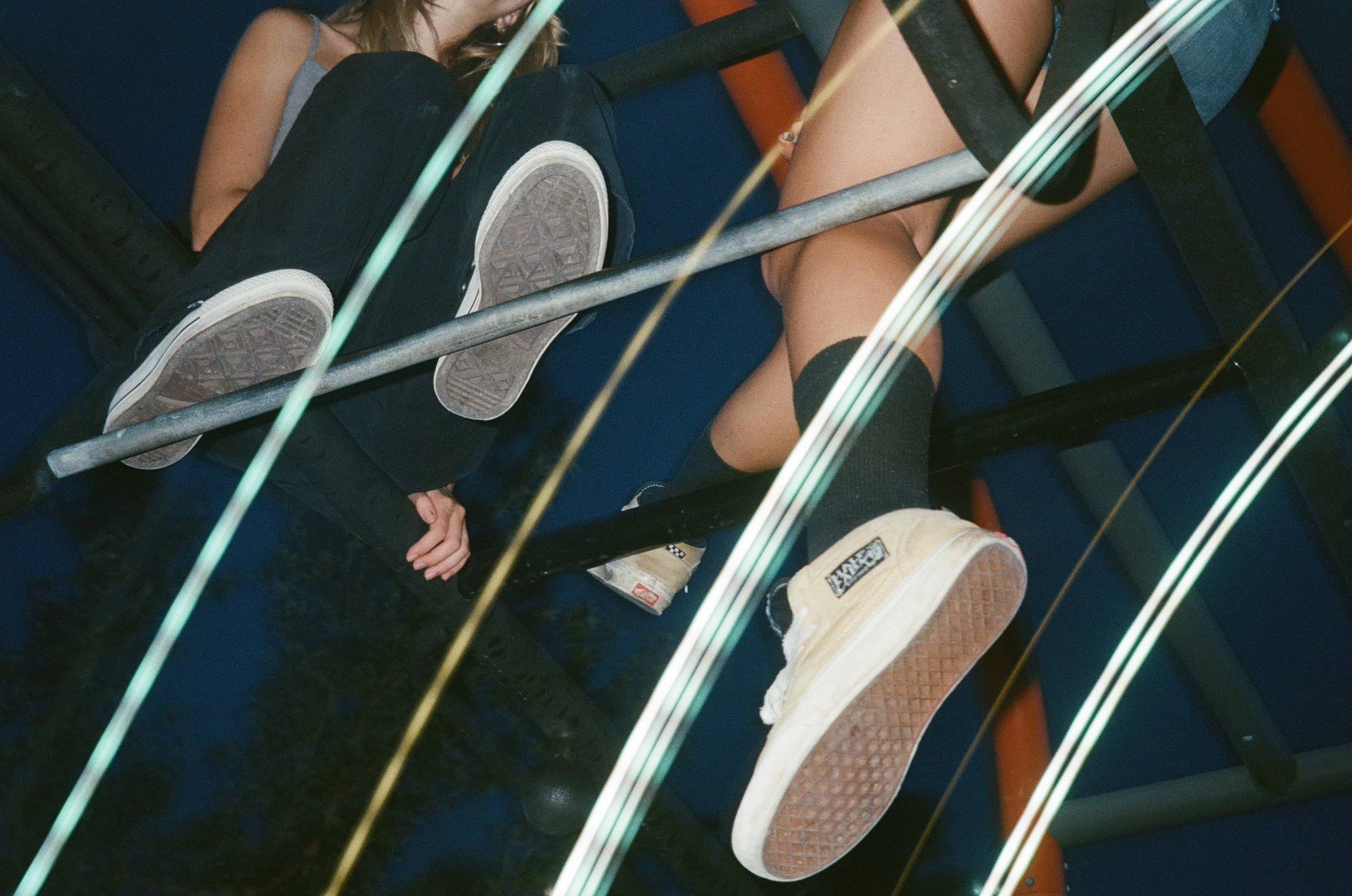 A person lying on a blue couch, wearing gray tank top and black pants, with their legs extended and wearing sneakers. The photo is taken through a glass railing with metallic bars and vertical reflective strips.
