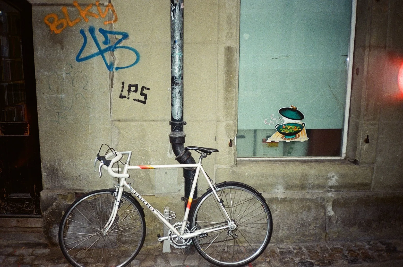 A white Peugeot bicycle leaning against a black pipe attached to a concrete wall with graffiti. There is a window to the right with a cartoon sticker of a steaming bowl of food on the glass.