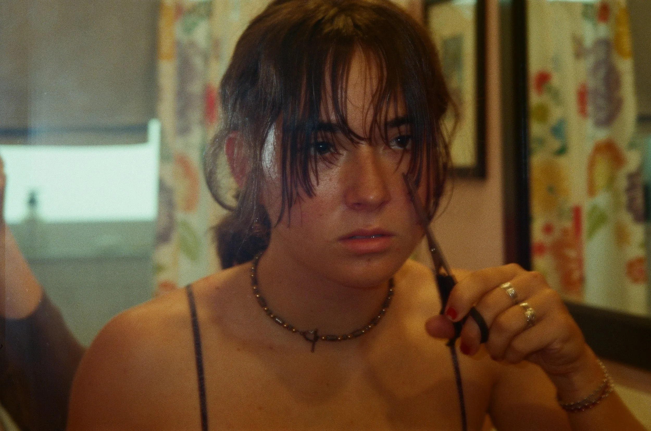 A woman with wet, messy hair looking into a mirror, wearing a spaghetti strap top, jewelry, and earrings, with a colorful background.
