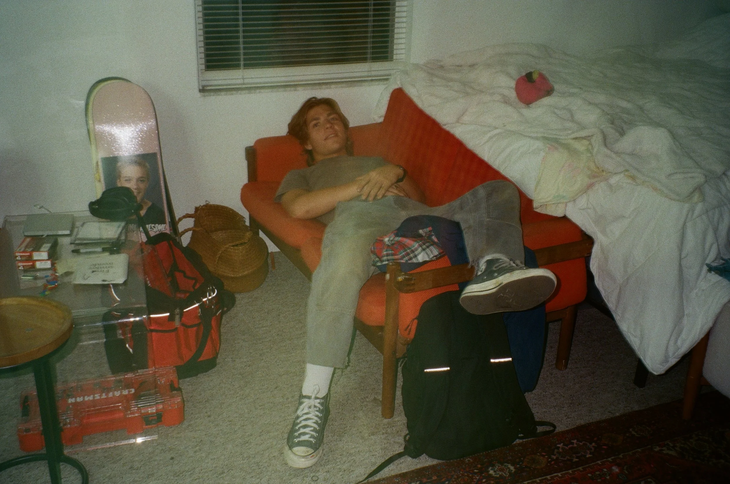 A young man with reddish-brown hair lying on a red couch in a bedroom, beside a bed with a stuffed animal on it. There is a table with various objects, including a mirror, magazines, and a red backpack, in the room.