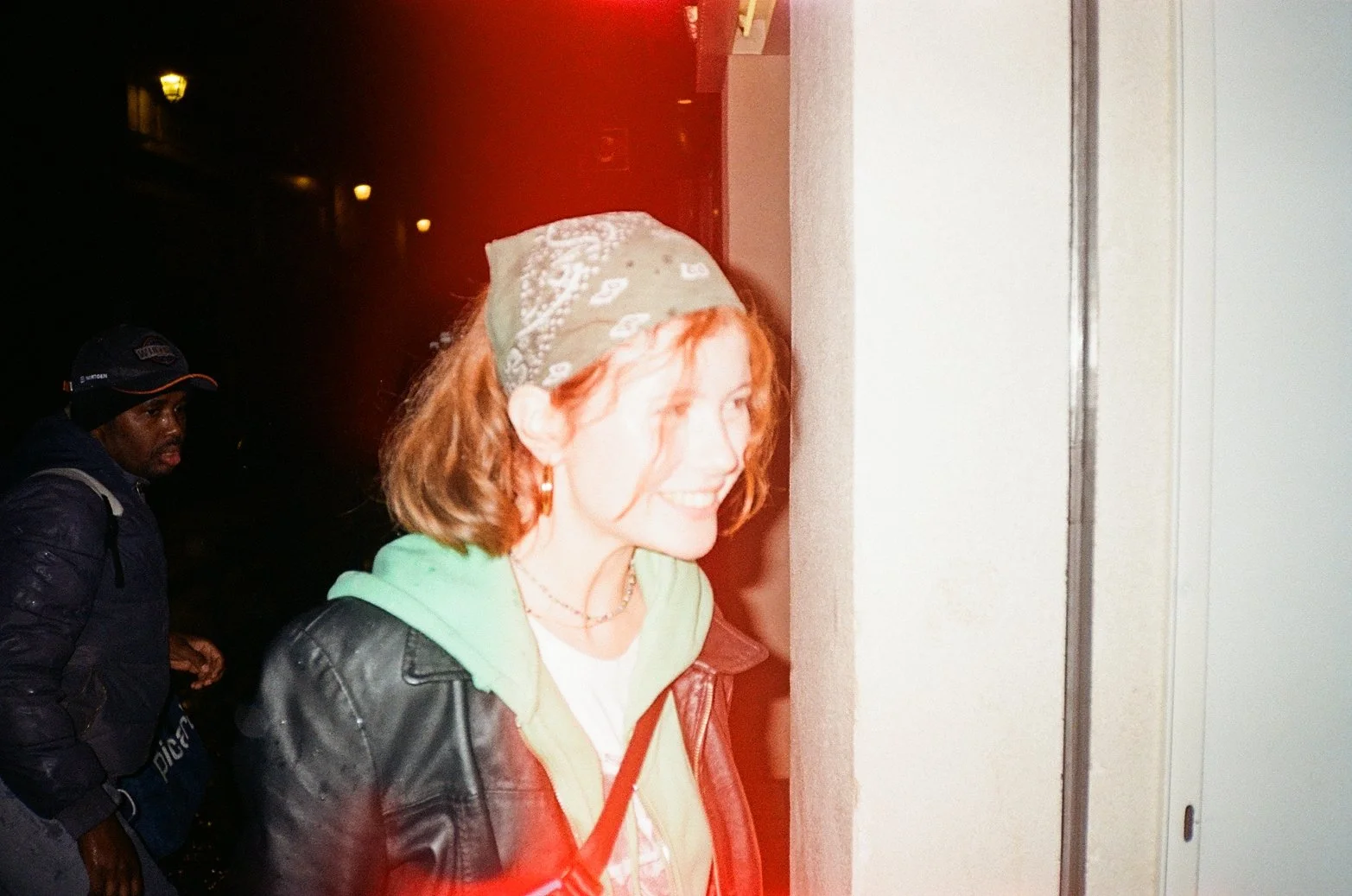 A woman with red hair, wearing a green hoodie, leather jacket, and a bandana, is smiling and looking at an elevator door. A man in a dark jacket and cap stands behind her, outside at night.