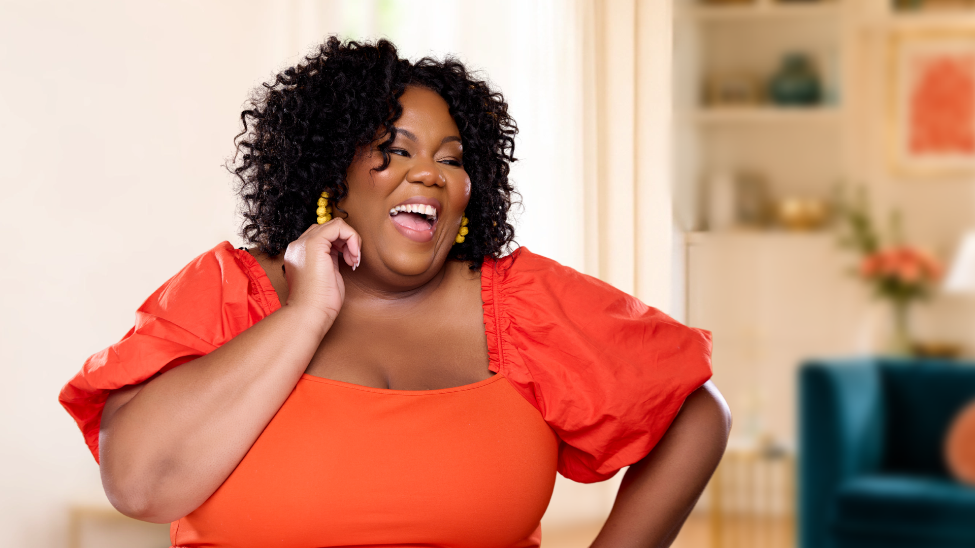 Amanda Ross-Hardin  woman wearing an orange top and yellow earrings, with curly black hair, is posing indoors with a blurred background of shelves, a framed picture, and a plant.