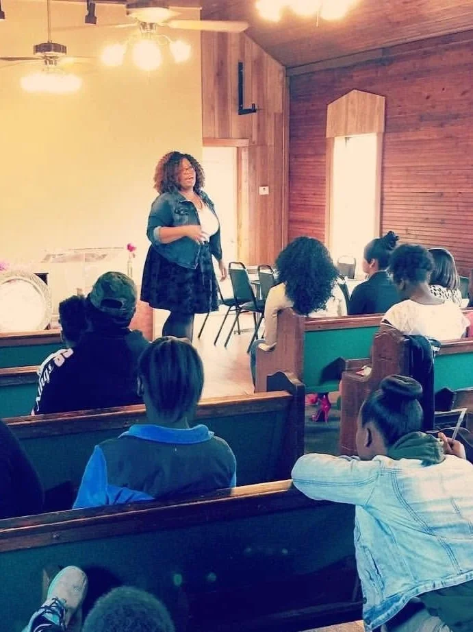 Amanda Ross-Hardin in front of a group of young women giving a presentation or speech. The children are sitting on benches, listening attentively.