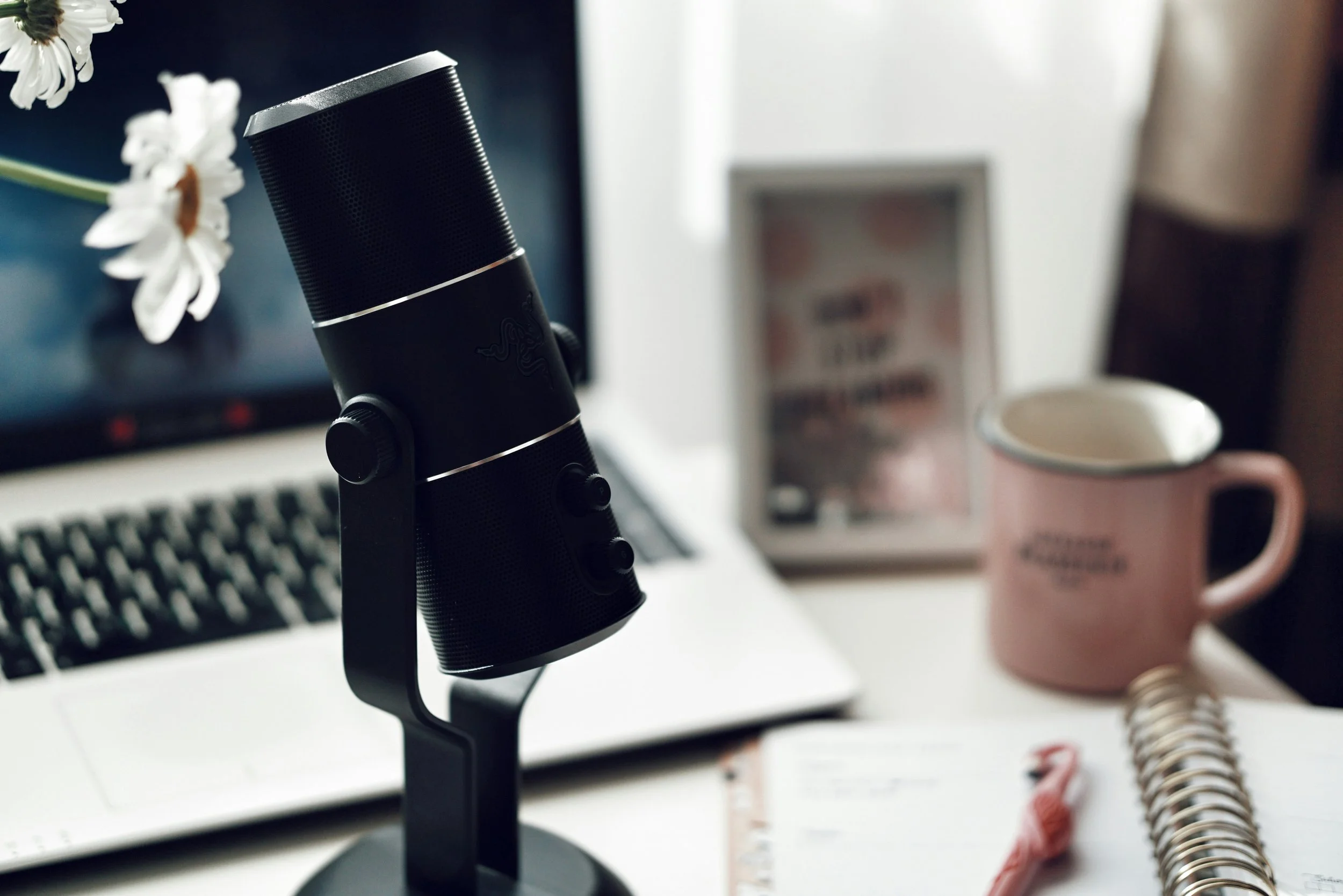 Close-up of a black microphone on a desk, with a computer keyboard, a framed paper, a pink mug, and a spiral notebook with a red pen in the background.