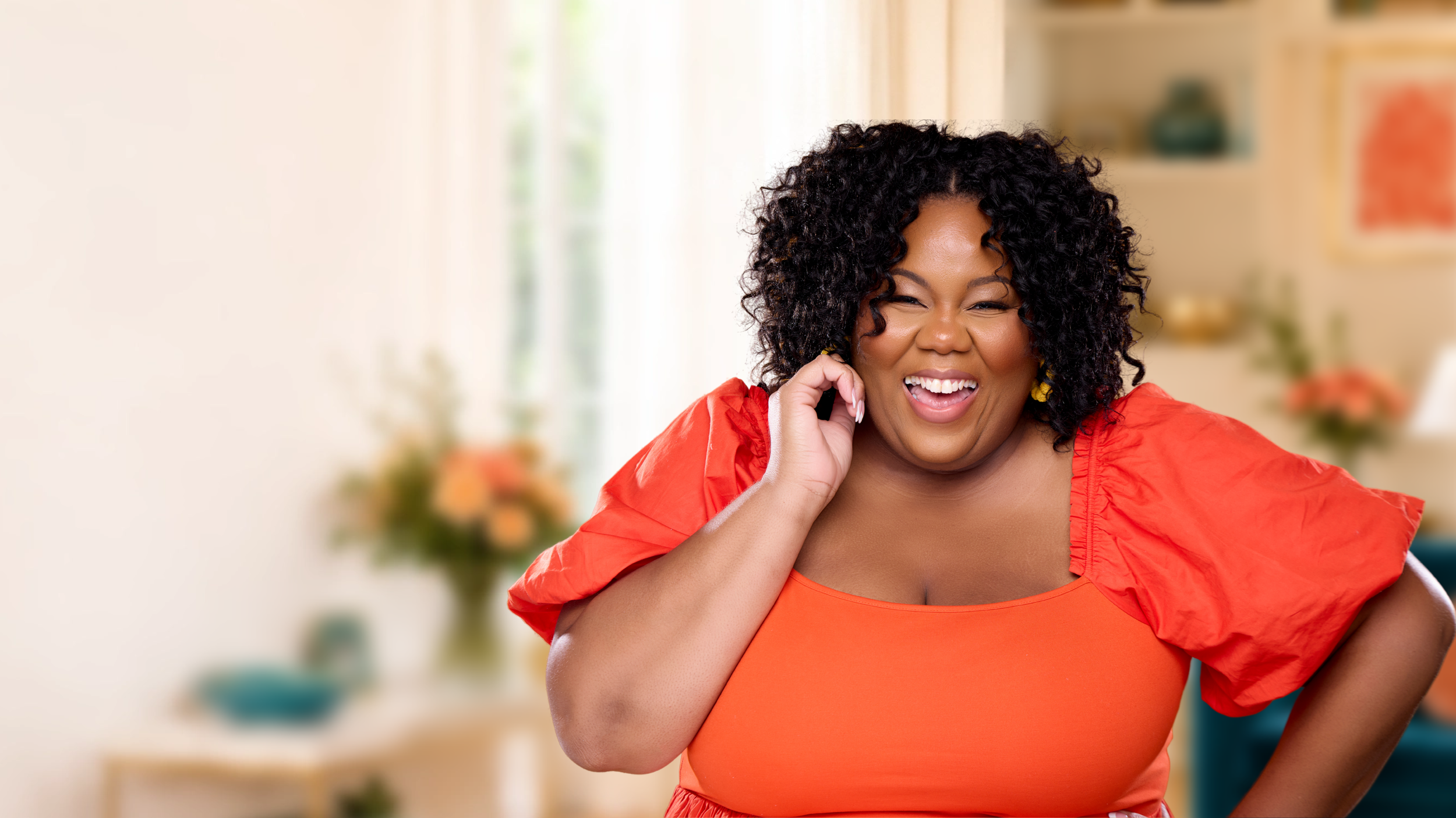 Amanda Ross-Hardin smiling and touching her face, wearing an orange dress, in a bright room with blurred background.