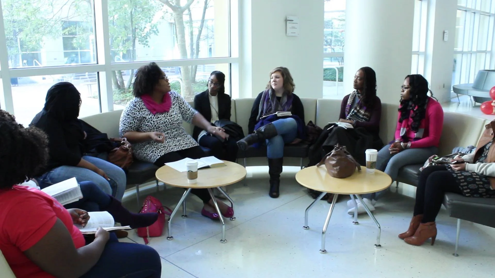 Amanda Ross-Hardin leading a group of women engaged in a discussion in a modern, well-lit room with large windows, small tables with coffee cups, and casual seating.