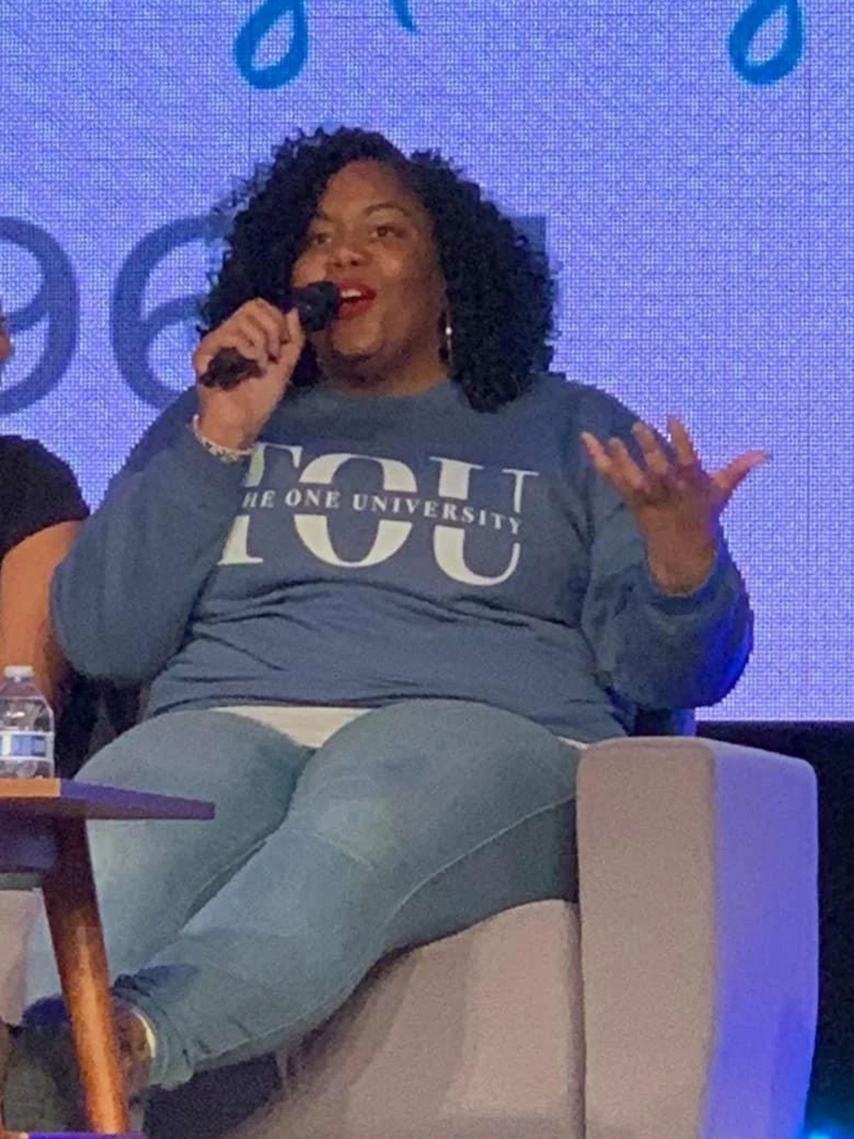 Amanda Ross-Hardin speaking into a microphone, sitting on a beige armchair, wearing a blue T-shirt with white text, in front of a blue patterned background.